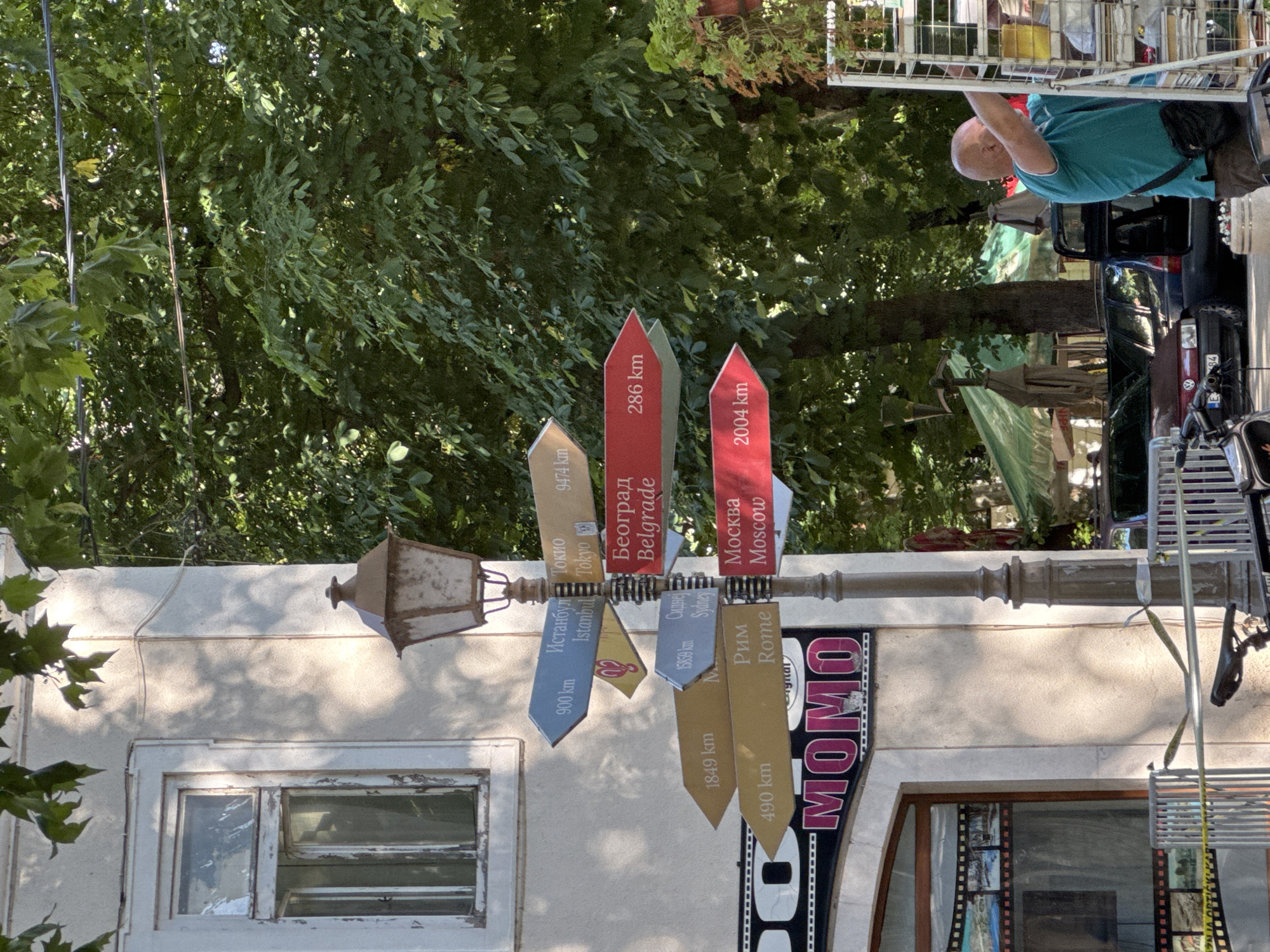 This media file depicts a street scene in Trebinje, Bosnia and Herzegovina, likely during daytime given the bright sunlight casting shadows. The primary subject is a directional signpost with multiple colorful arrows pointing to various cities and their distances, including Istanbul, Tokyo, Belgrade, Moscow, Sydney, and Rome. Text on the arrows is in both Cyrillic and Latin scripts, indicating international destinations.
In the background, lush green trees dominate the upper portion of the frame, suggesting a warm season. A building with a light-colored, textured facade and a window with visible panes is visible on the left. A vintage-looking lamp hangs from the signpost. A person, a bald man wearing a teal shirt and dark pants, is partially visible in the upper right, interacting with a metal structure that could be part of a market stall or display. A car is parked behind him. Below the signpost, a prominent sign reads "MOMO" in large, pink letters, likely indicating a business or establishment. The overall scene suggests a lively, perhaps tourist-oriented, urban environment. The presence of directional signs to distant global cities hints at Trebinje's role as a point of connection or a place with a global outlook.