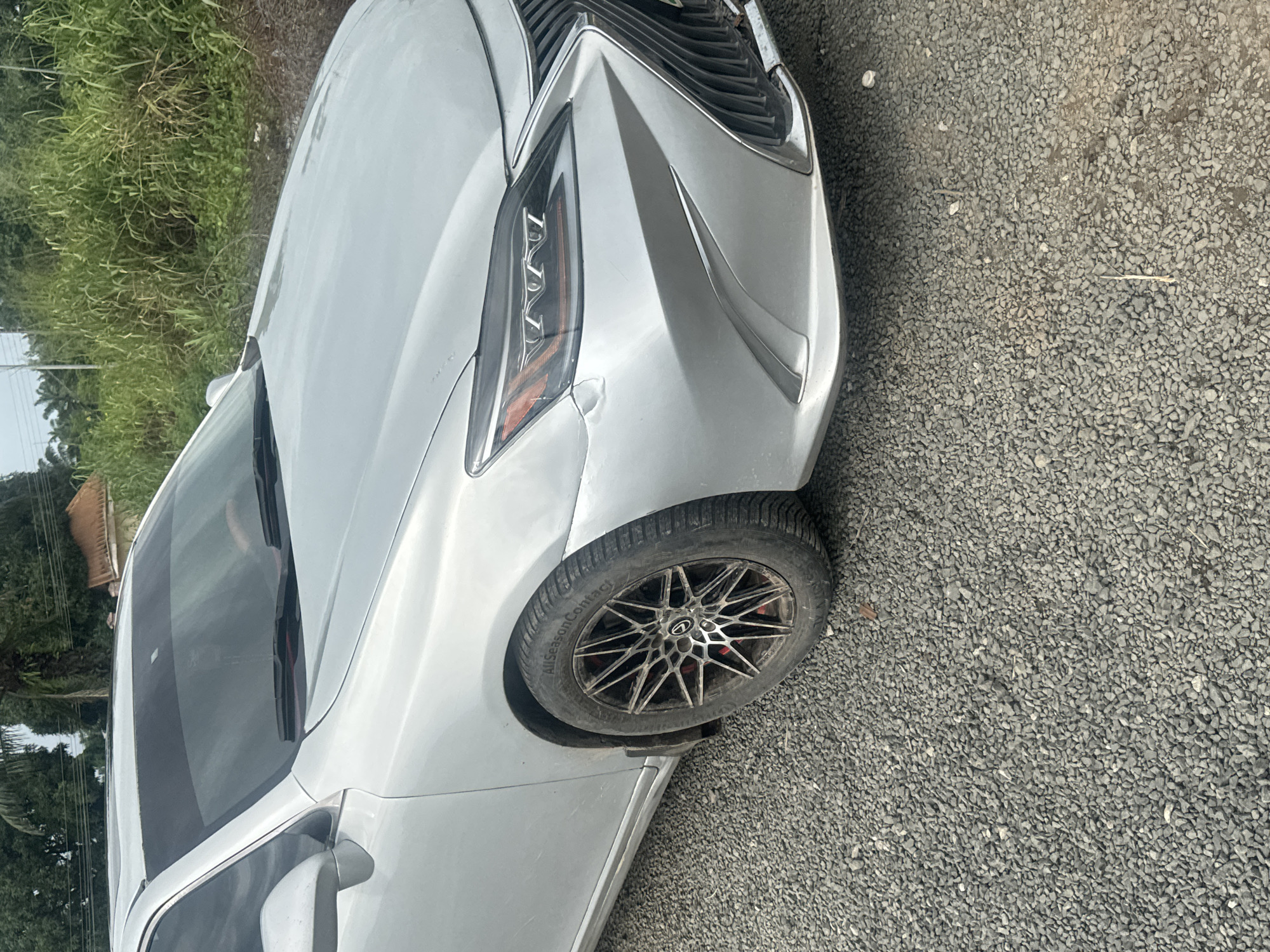 A silver sedan is parked on a gravel surface. The car's front left tire and fender are visible. The car has custom rims and the tire has "AllSeasonContact" written on it. In the background, there is dense green vegetation and a building with a brown roof. The scene appears to be in a rural or semi-rural area. There are no people visible in the image. The car appears to be in good condition, with no visible damage. The lighting suggests it is daytime.