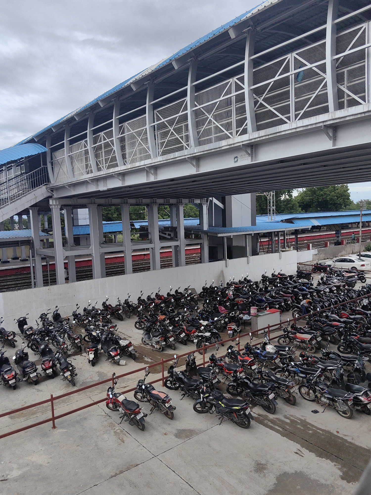 The image shows a large parking lot filled with motorcycles and scooters under a skywalk at the Malkapur railway station in India. The skywalk, made of grey metal and featuring blue roofing, spans across the scene, with its supports forming part of the structure. Below the skywalk, train tracks are visible, indicating the station's context. A substantial number of two-wheeled vehicles are parked tightly together, filling the paved area. Among the vehicles, a few people are visible, including one person walking near the train tracks and others in the parking lot. A white car is also parked in the background. The environment appears to be an outdoor public space, likely a transportation hub, with a mix of urban infrastructure and a natural green backdrop of trees in the distance.