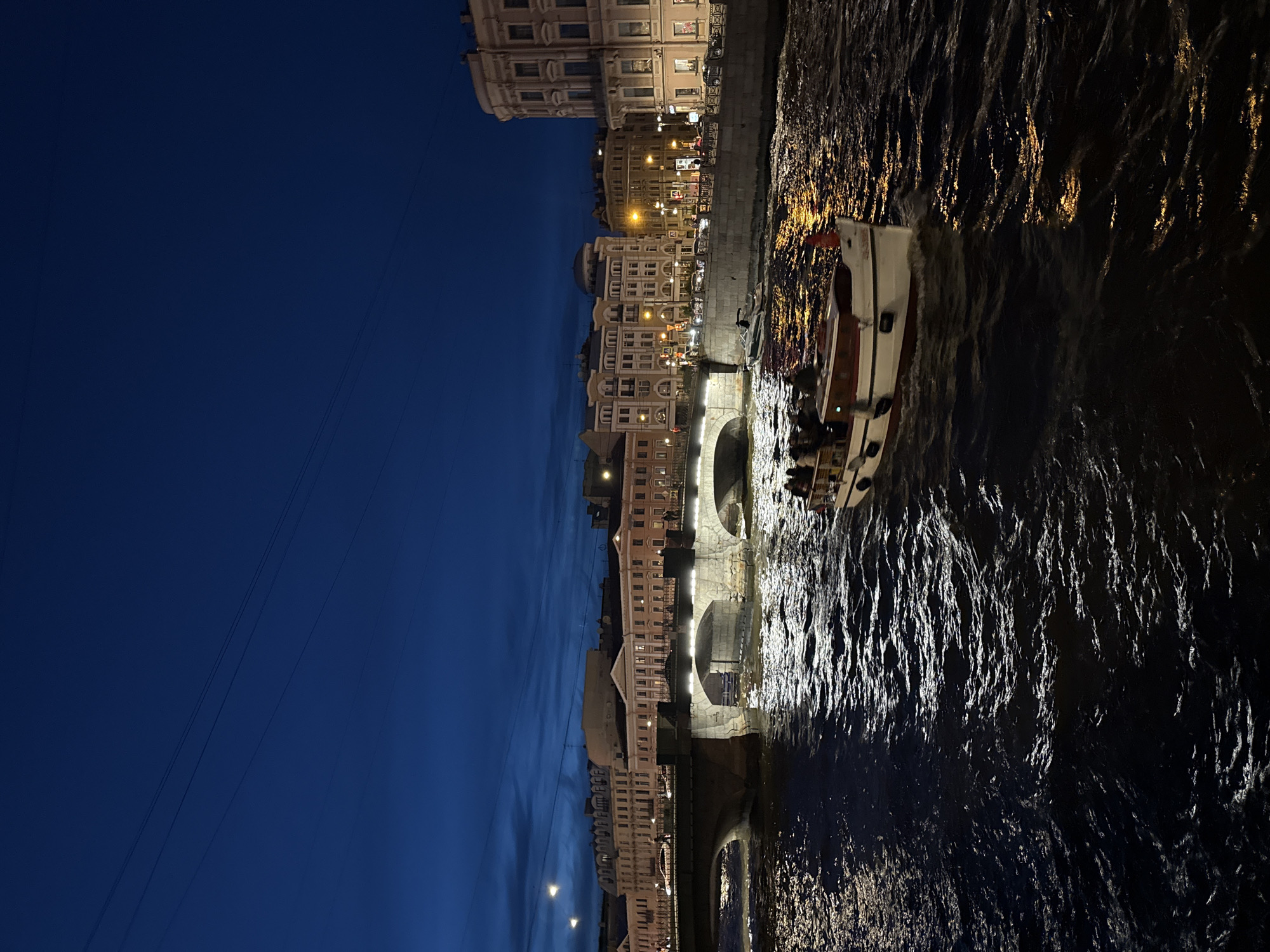 The image depicts a nighttime scene in Saint Petersburg, Russia. A tour boat carrying passengers is navigating the canal beneath the ornate bridge. The city’s architecture is illuminated along the canal's edge, with multiple buildings and lights visible. The sky is a deep blue hue. The water reflects the lights of the city, creating a shimmering effect.