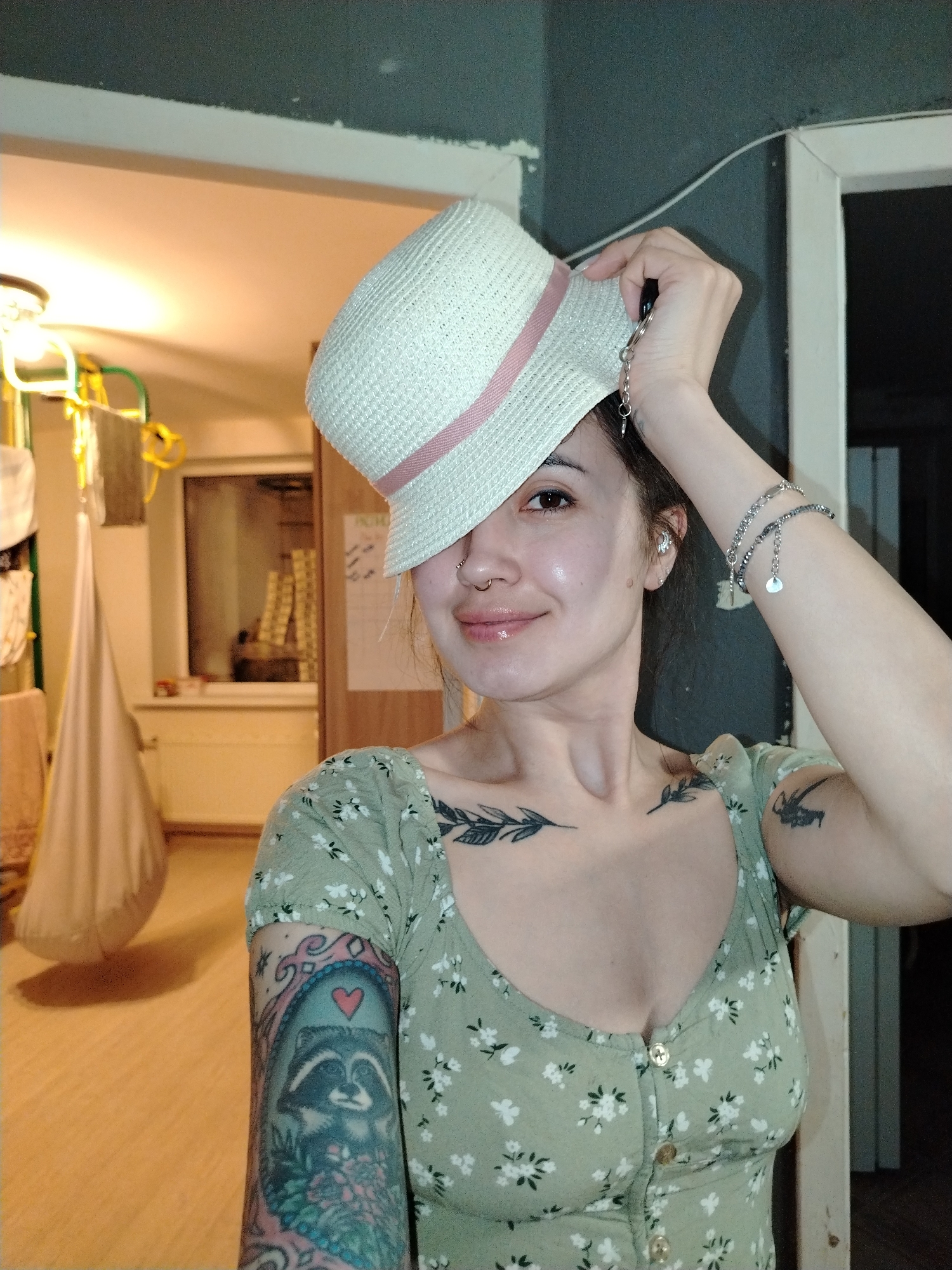 A woman is indoors, holding a white hat with a pink ribbon on her head, partially obscuring her left eye. She is wearing a light green floral print dress with buttons. She has a nose ring and multiple bracelets on her raised right arm. There are tattoos visible on her chest and left arm, including one of a raccoon in a frame. In the background, there is a doorway leading to another room with a clothes drying rack and hanging items. The room appears to be in a residential setting. The image does not contain information to determine whether the location is Noviy, Russia.
