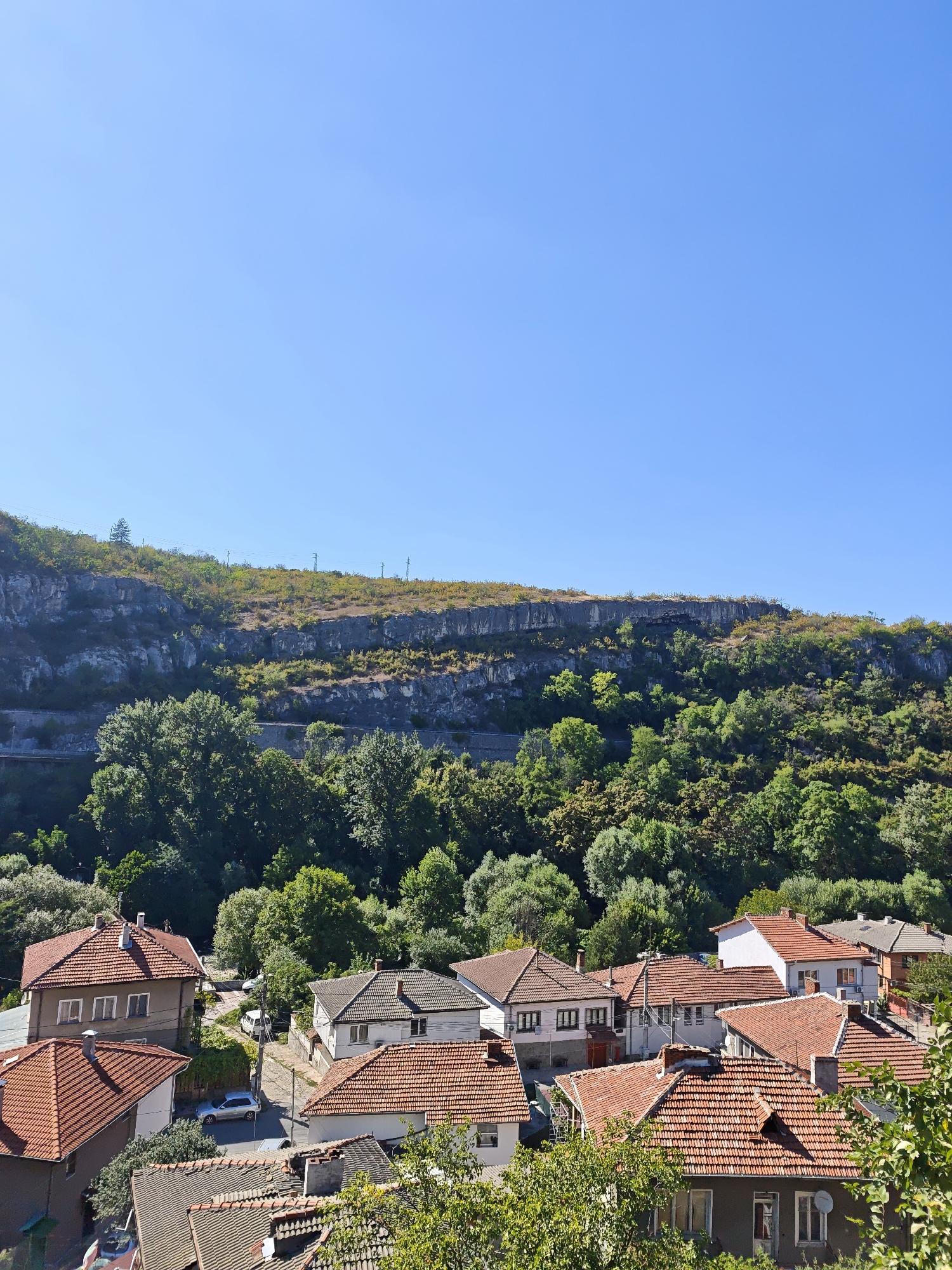 The image shows a view of a residential area with houses featuring red tile roofs, nestled at the foot of a steep, rocky cliff face covered in lush green trees. The sky is a clear, vibrant blue, indicating a sunny day. In the foreground, several houses are visible, showcasing typical Balkan architecture with their tiled roofs and light-colored walls. Some of the houses have multiple windows and chimneys. A few cars are parked on a paved road that winds through the neighborhood. In the background, the imposing cliff rises dramatically, its rugged surface dotted with vegetation. The overall scene depicts a tranquil village or small town setting, characteristic of regions like Lovech, Bulgaria, known for its picturesque natural landscapes and historic architecture.