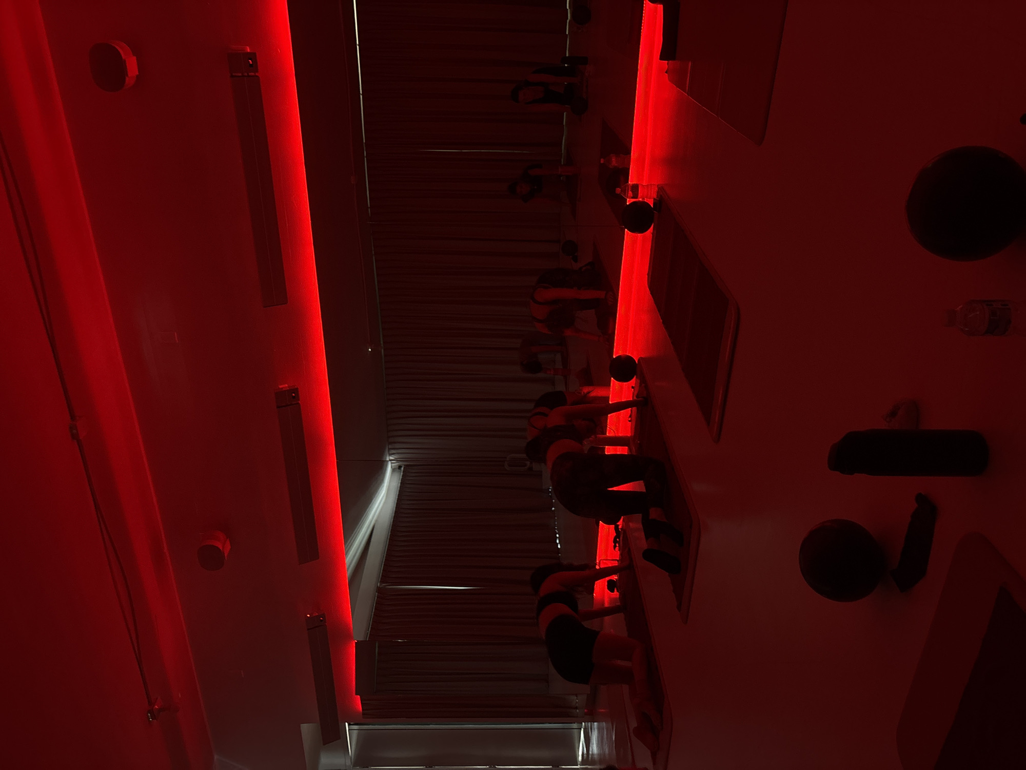 The image depicts a workout class in a room illuminated with red light, likely infrared. The location is a hot yoga studio in Mexico City, Mexico. Several people, presumably women, are aligned on yoga mats, participating in a fitness routine that involves bending or stretching. Water bottles and workout equipment are visible. The environment has a contemporary design, with linear lights and a minimalistic aesthetic.
