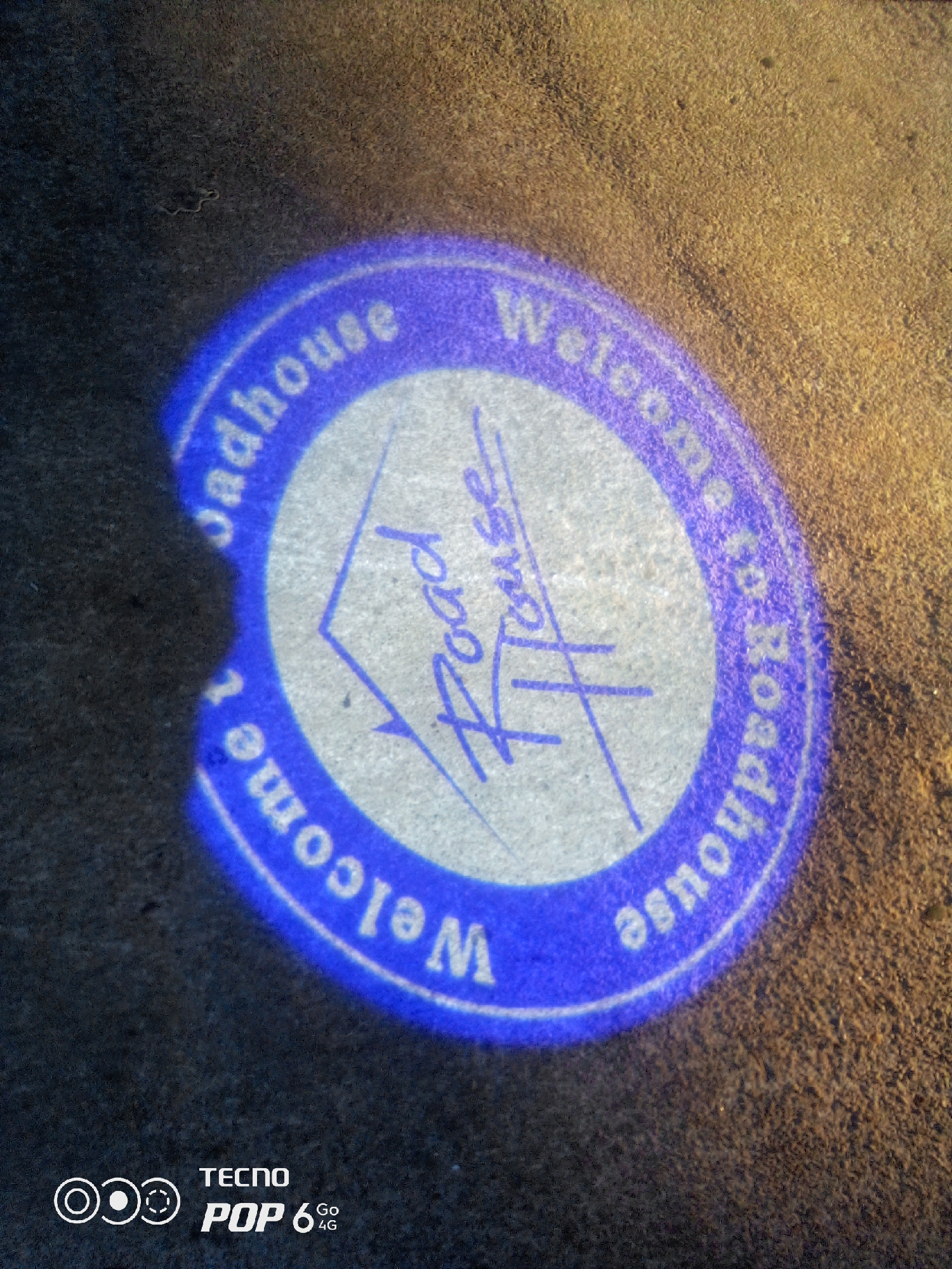 An illuminated logo projected onto a gritty, sandy surface reads "Welcome to Roadhouse". The logo is circular with a blue outer ring and a white inner circle. Within the inner circle, the words "Welcome to" arc around the top and right side, while "Roadhouse" appears in script lettering in the center, slightly tilted. The words are white and appear to be illuminated by a blue light source, causing a strong blue glow around the text. The sandy surface is textured and appears to be outdoors. In the bottom left corner of the image, the TECNO POP 6 4G logo is visible. The overall scene suggests an entrance or walkway illuminated by a branded projection.