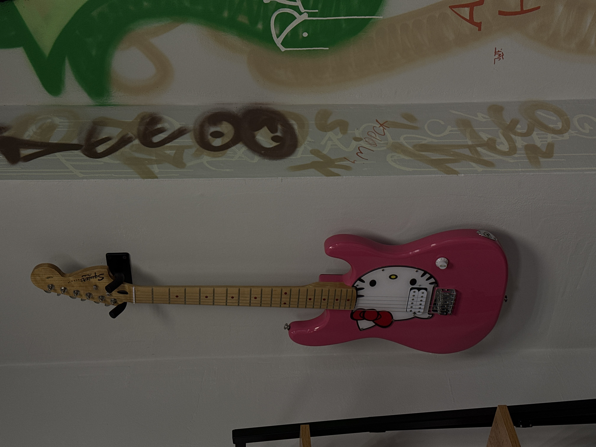 A pink Hello Kitty electric guitar is mounted on a black wall mount against a white wall. The guitar has a wooden neck and fretboard with red fret markers. The body of the guitar features a Hello Kitty graphic with black eyes, a yellow nose, and a red bow. To the right of the guitar, a concrete pillar is covered in graffiti. The graffiti includes brown and light brown spray-painted tags and symbols, as well as a large green abstract shape. A small red signature, "J.W.F.", is visible on the pillar. A portion of a black metal shelf unit is visible to the left of the guitar.