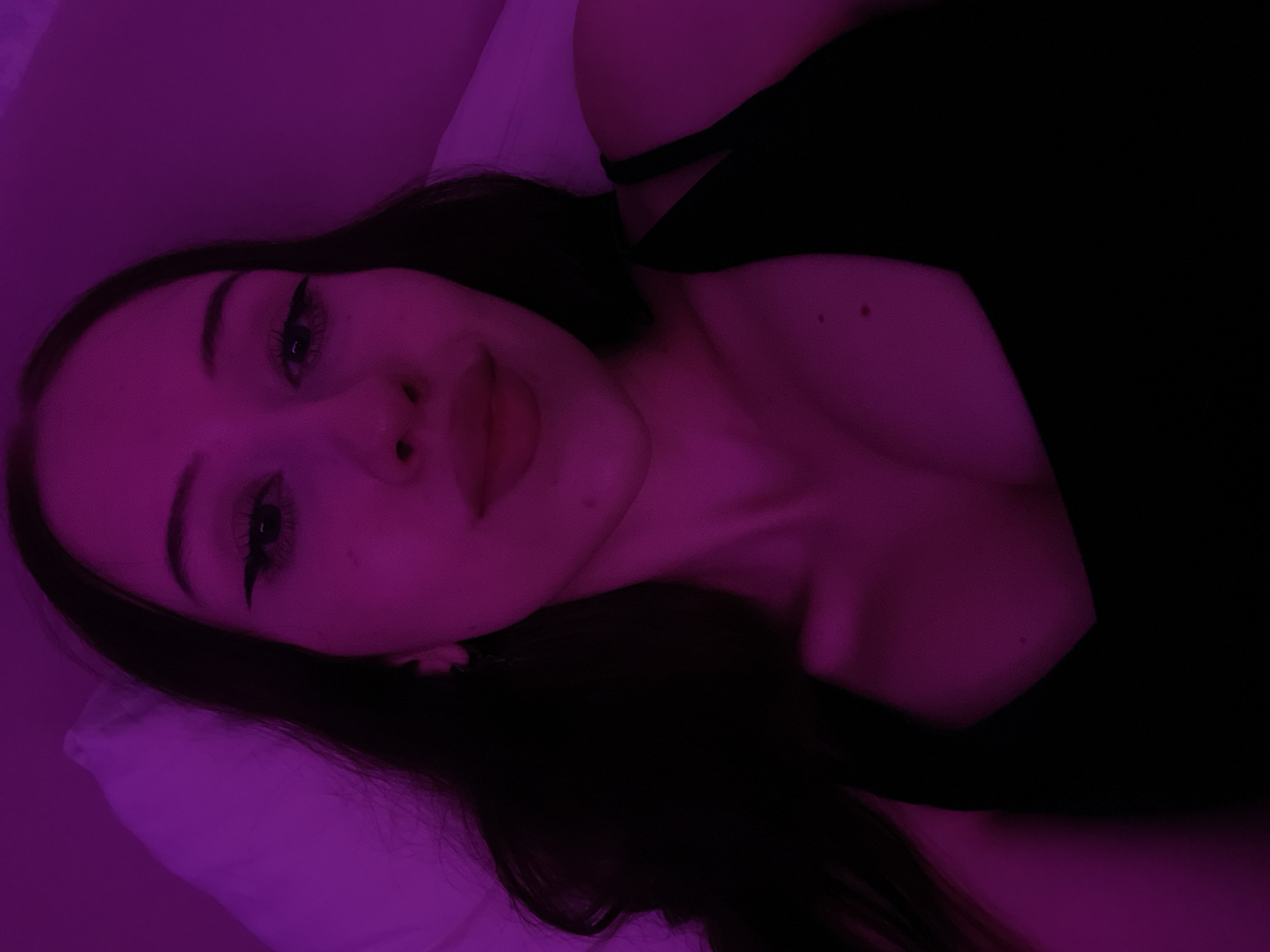A young woman with long, dark hair and winged eyeliner lies on a pillow, her head tilted back. She is wearing a black tank top and is illuminated by a vibrant pink light. The background is a blurred pink wall and a white pillow. No specific location within Votkinsk, Russia, can be identified.