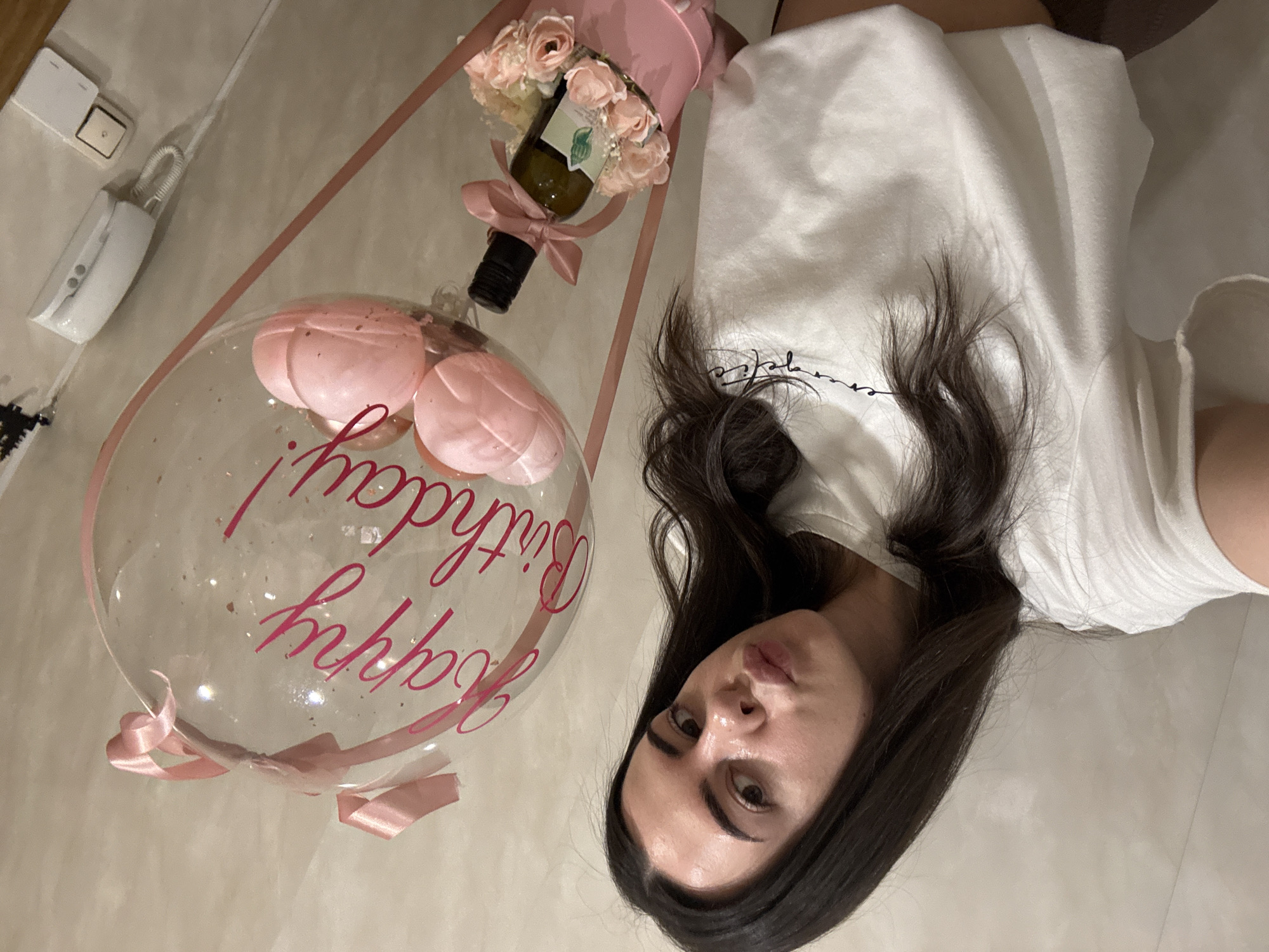 A young adult female with long, dark, wavy hair, dark eyebrows, and pursed lips is depicted in a selfie, looking directly at the camera. She wears a white, short-sleeved t-shirt with dark script lettering, partially visible, on the chest. She holds a birthday gift consisting of a clear, spherical balloon inscribed with "Happy Birthday!" in hot pink script. The balloon contains small, pink, ribbed spherical objects and gold-colored glitter. Two pink ribbons are tied at the top of the balloon and descend to a cylindrical pink box at the bottom, creating a hot-air balloon aesthetic. The pink box holds a small, dark-colored bottle with a light green label and a pink ribbon tied around its neck, surrounded by light pink artificial roses. The background features a light beige, textured wall. On the right side of the wall, a white intercom unit with a coiled cord and two buttons is mounted, along with a light-colored electrical outlet or switch plate positioned below it. A dark, possibly stylized tree or abstract shape, is affixed to the wall above the gift. The scene is indoors in Unknown, Unknown.