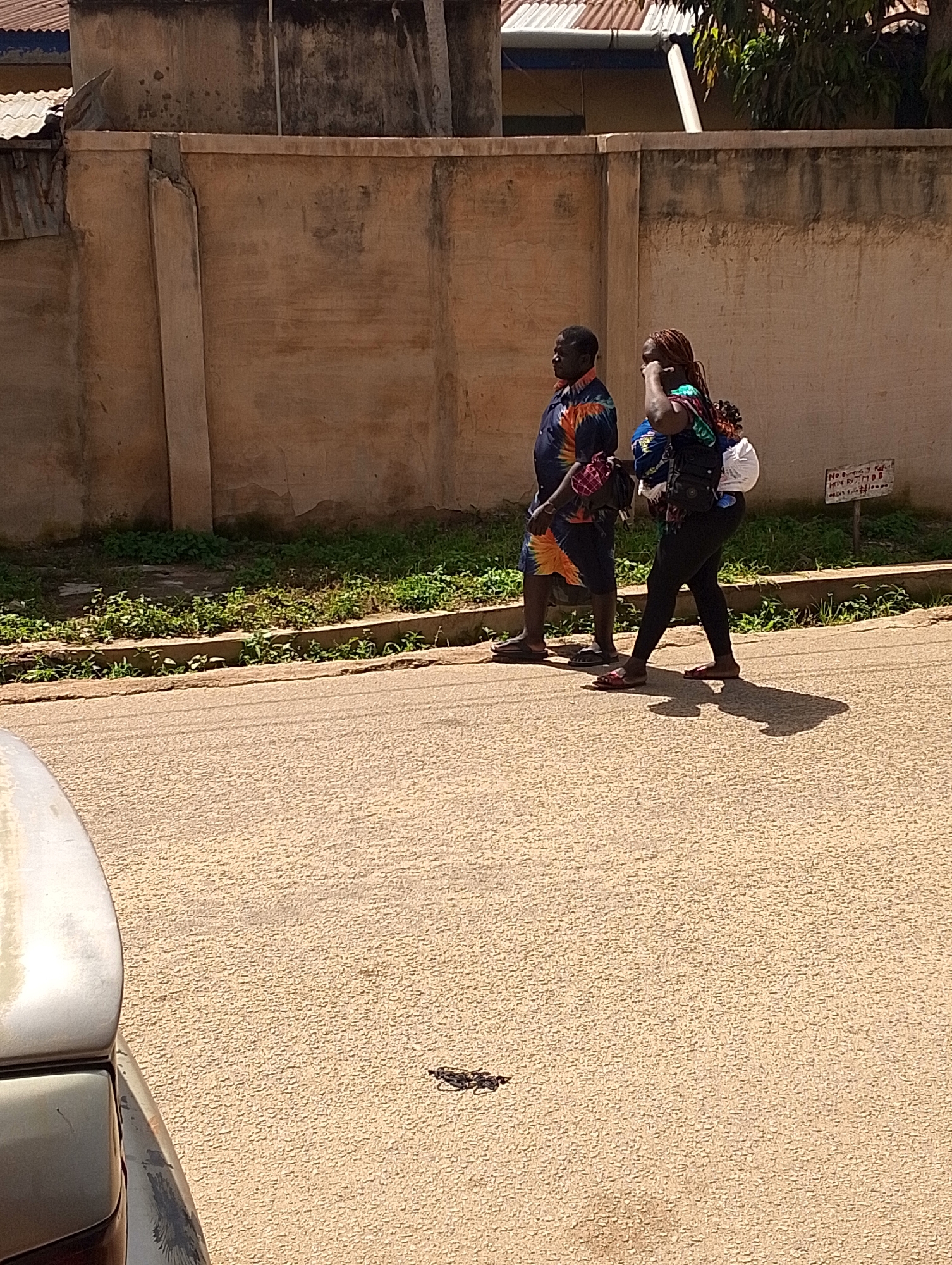 The image shows two people walking on a paved road. The person on the left is a man wearing a colorful, short-sleeved shirt and dark shorts. He is wearing dark sandals. The person on the right is a woman with braided hair, wearing a dark top and black leggings. She is carrying several items wrapped in fabric on her back and appears to be wearing sandals. They are walking side-by-side, and their shadows are cast on the road, suggesting it is daytime, likely mid-morning or afternoon, with bright sunlight.
They are walking past a low concrete wall with a dirt and grass embankment behind it. Beyond the wall, there is a building with a corrugated metal roof and some trees. The wall is weathered and appears to be part of a property boundary. There is a small, weathered wooden sign on a post to the right of the figures, attached to the wall. It contains some handwritten text, which is partially legible and appears to be in English, possibly indicating a price or information about goods or services. The road surface is asphalt with visible texture. A portion of a car's hood is visible in the lower left corner of the frame, suggesting the photo was taken from a vehicle or from a low vantage point near the road. The overall scene depicts a common street view in a populated area.