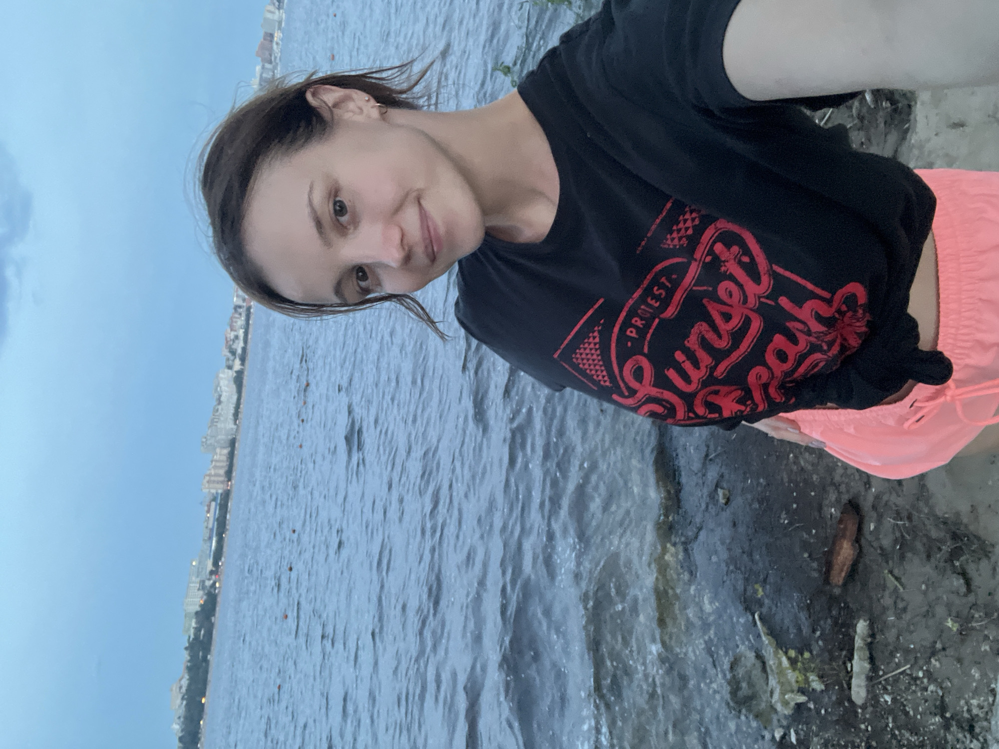 A young woman is taking a selfie by the water. She is wearing a black t-shirt with a red graphic and bright pink shorts. She has dark hair pulled back and is smiling at the camera. In the background, there is a wide body of water with a city skyline visible on the opposite shore. The sky is overcast, suggesting it is either early morning or late evening. The ground she is standing on appears to be a sandy or muddy shore with some debris, including driftwood. The image was taken outdoors, likely near the Volga River, given the city skyline.