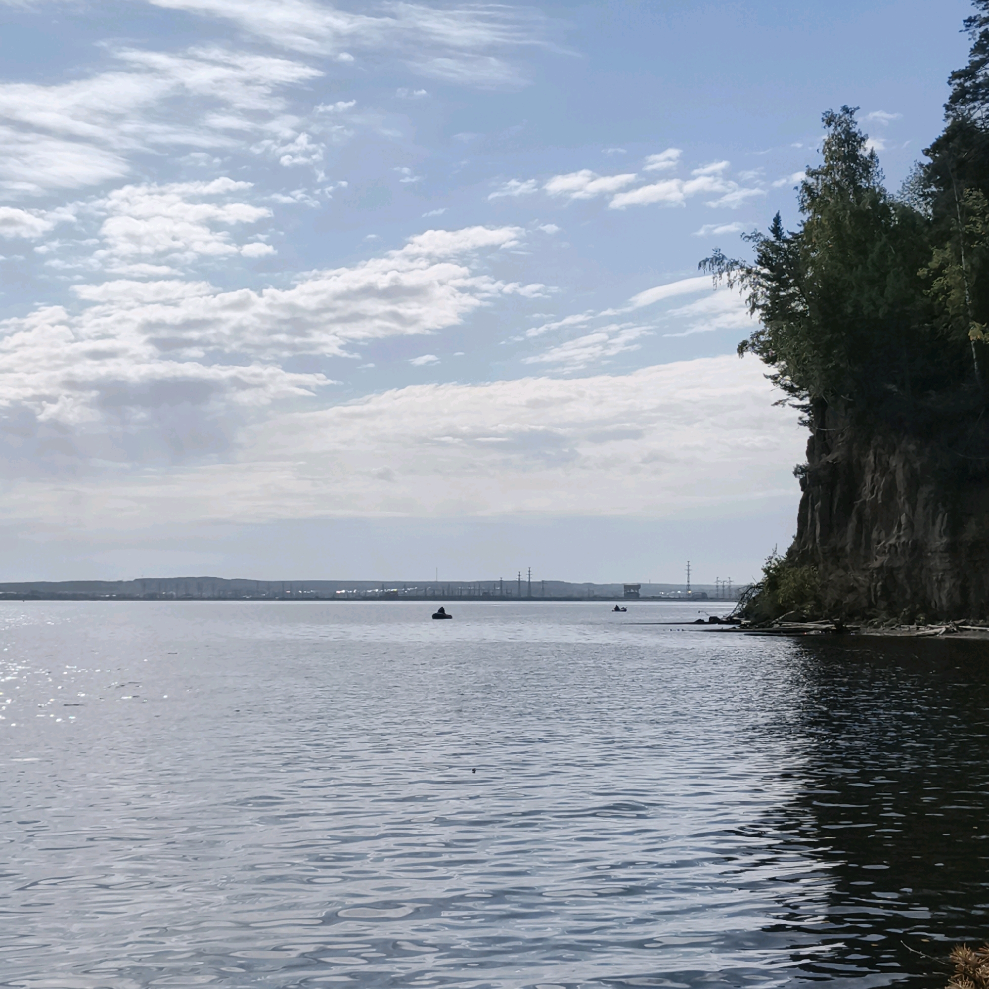 This image depicts a wide, calm body of water, likely a river or reservoir, with a steep, rocky cliff face covered in dense green foliage on the right side. The sky above is a pale blue with scattered white clouds. In the distance, along the opposite shore, a faint industrial skyline is visible, characterized by tall structures and power lines. There are a few small boats or kayaks on the water. The scene appears to be set in or near the city of Chaikovsky, Russia, possibly along the Kama River. The lighting suggests it is daytime, with the sun casting a slight sheen on the water's surface. The overall atmosphere is serene and natural, juxtaposed with the distant signs of industrial activity.