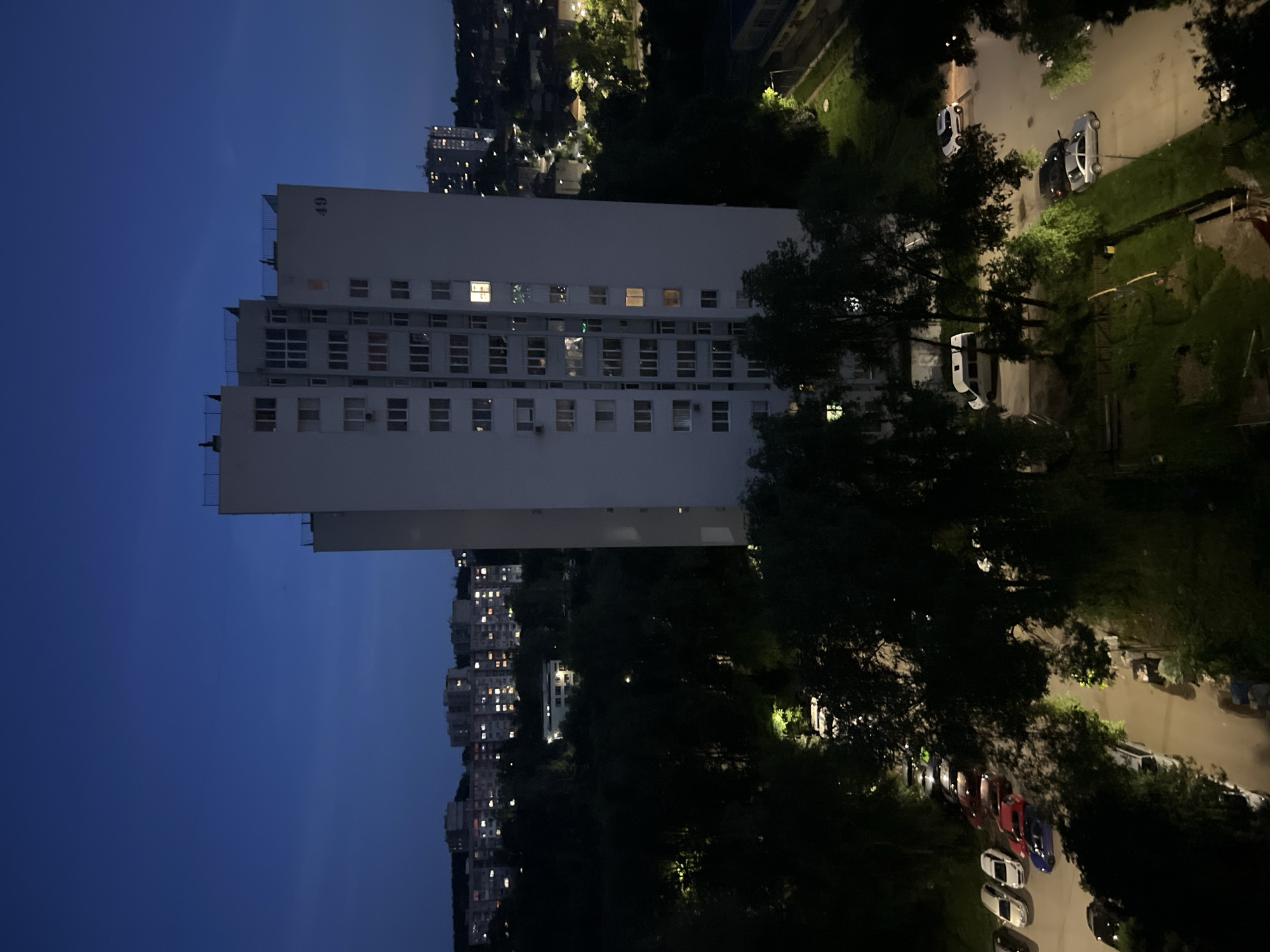 The image shows a high-rise apartment building at dusk, located in the city of Dobrich, Bulgaria. The building, numbered "49", is the most prominent structure in the foreground. Several other buildings, characteristic of residential areas, are visible in the background, with lights on in some of the windows, suggesting it is evening. The surrounding area is covered with trees and a parking lot with several cars. The sky is dark blue, indicating the transition from daylight to night.