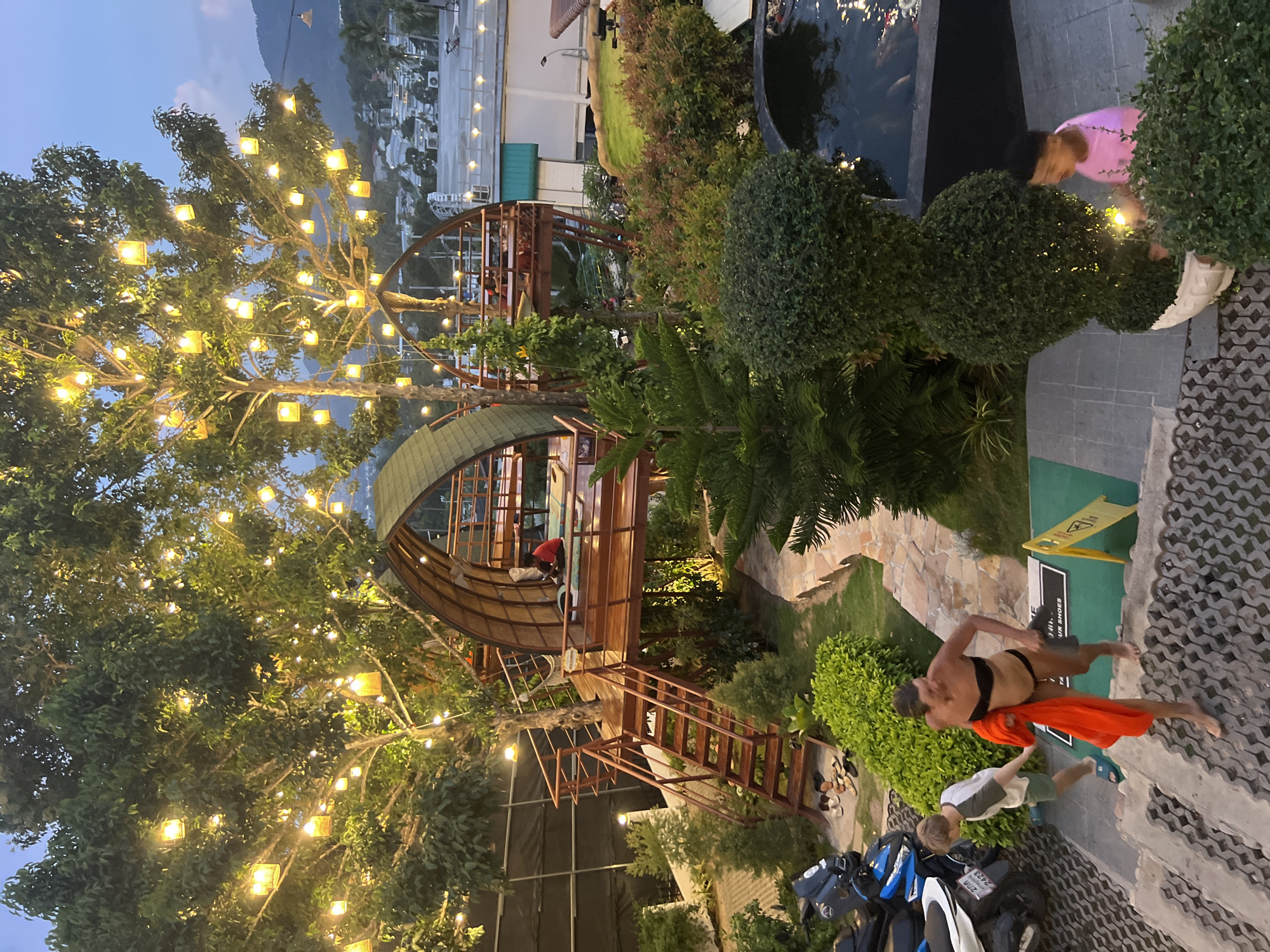 The image depicts a scenic outdoor dining area, likely a restaurant or resort, in the city of Ko Samui District, Thailand, as evidenced by the tropical vegetation and mountainous backdrop. The setting is enhanced by numerous decorative lights strung across a large tree, creating a warm and inviting ambiance.
In the foreground, two people are descending a stone staircase. A young boy, wearing a white t-shirt and green shorts, walks ahead, while a woman, dressed in a black bikini and carrying an orange towel, follows closely behind. To the left, several motorcycles are parked, suggesting guests have arrived by vehicle.
A unique architectural feature is a raised wooden structure resembling a treehouse, with a curved roof and open-air seating. A person in a red shirt is visible sitting inside this structure. The area is densely landscaped with various trees and bushes, and a koi pond is visible in the lower right portion of the image. The overall atmosphere suggests a tranquil evening setting with a touch of rustic charm.