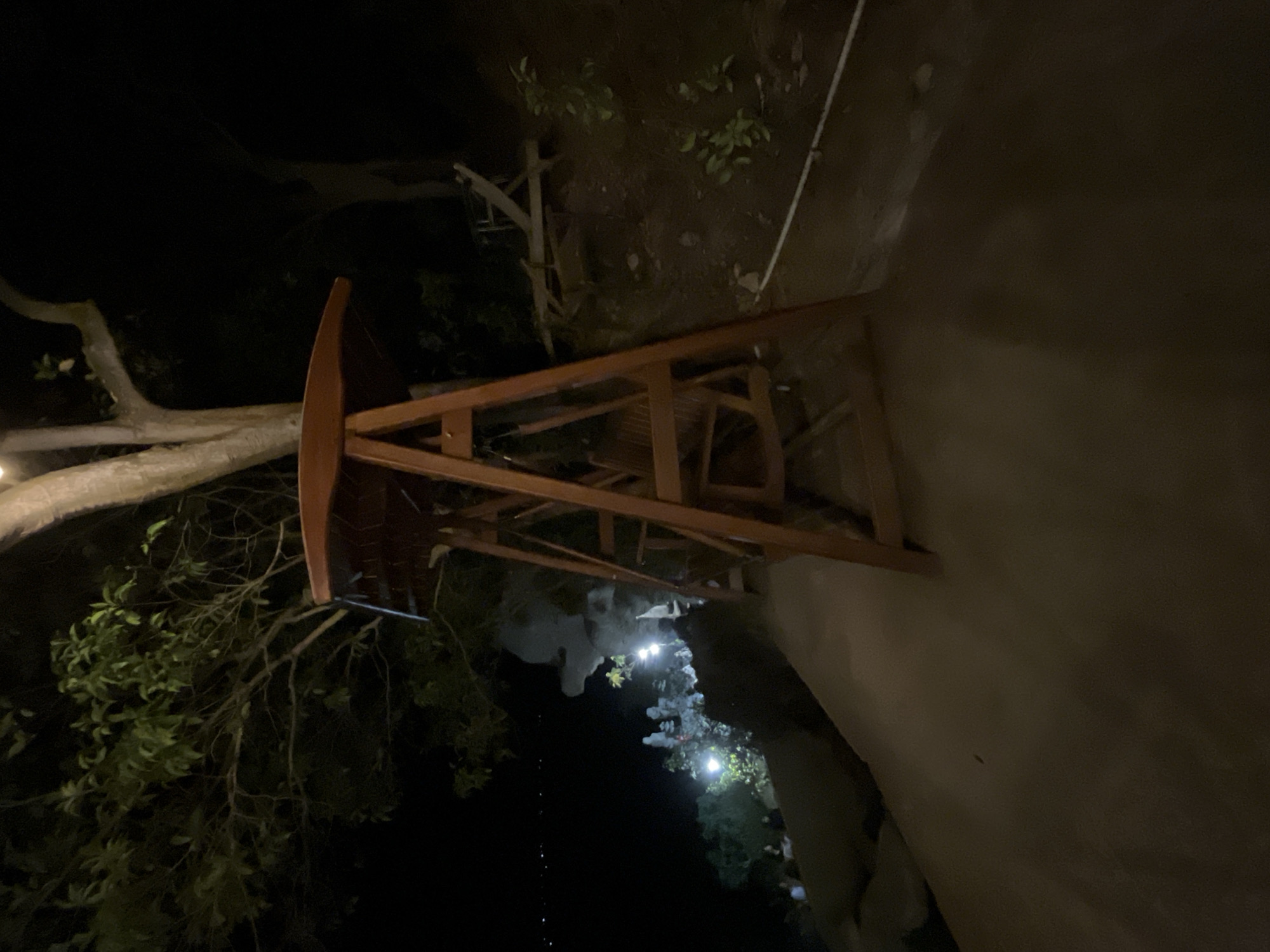 The image captures a night scene at a location in Karimunjawa, Indonesia, specifically on a pathway leading to a vantage point overlooking a body of water. In the foreground, a wooden swing set stands prominently, its frame and seat made of dark wood. Behind the swing, a large tree with visible branches and foliage extends upwards, partially illuminated. To the left of the swing, a steep embankment slopes down towards the water, where a cluster of people can be seen gathered. The water itself is dark, reflecting distant lights from what appears to be a shoreline or boat activity. The ground is a dark, uneven surface, possibly a dirt or gravel path. The overall atmosphere is dimly lit, suggesting it is nighttime, with artificial lights from the gathering of people and possibly some ambient lighting contributing to the scene.