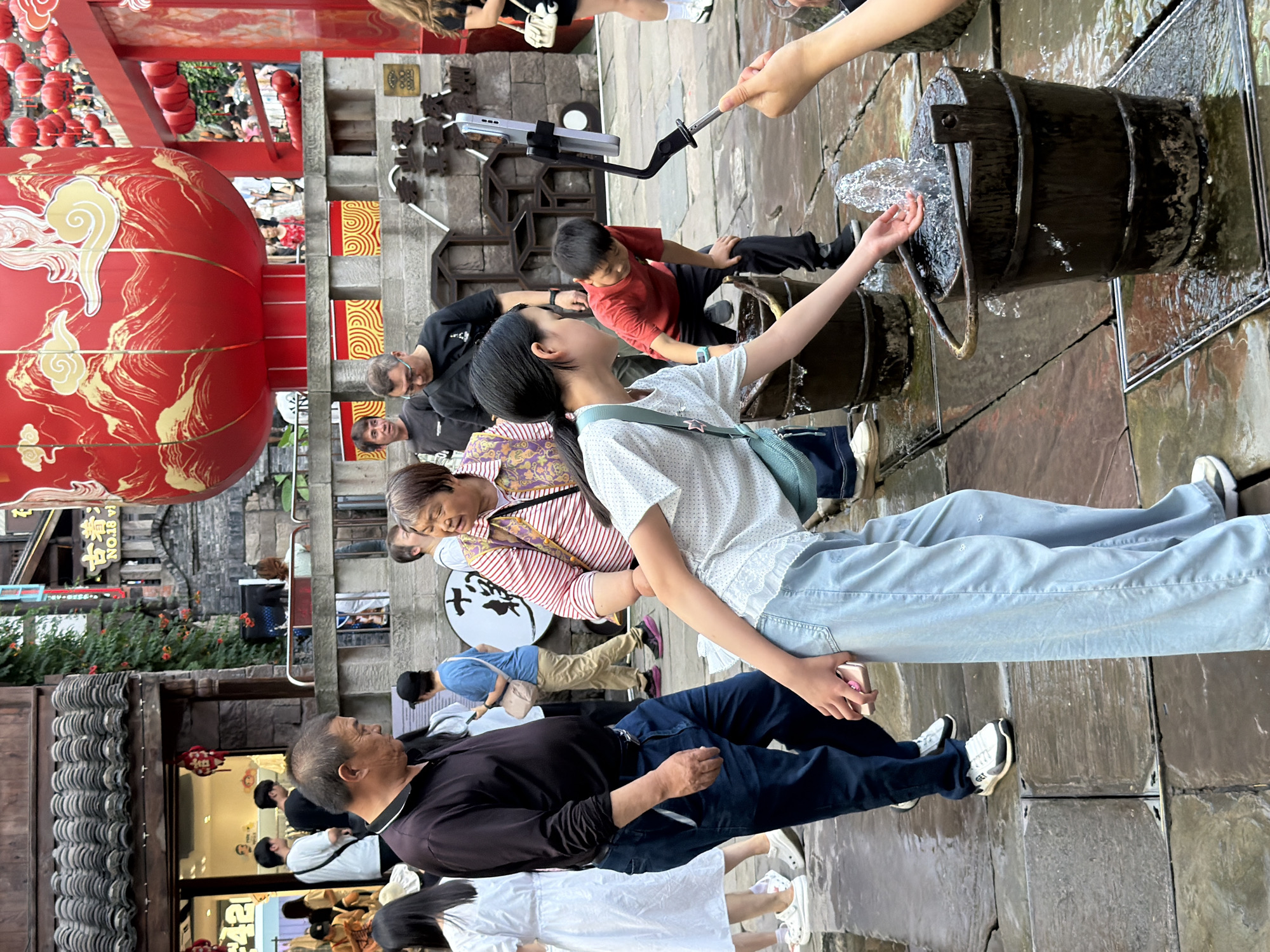 The image captures a street scene in the Yuzhong District of China, characterized by traditional architecture with tiled roofs and stone walls. Dominating the background is a large, ornate red lantern, a common festive decoration. In the foreground, several people are engaged in various activities. A young girl in a white patterned shirt and light blue pants is interacting with a fountain system, with water splashing from a bucket. She is holding a pink smartphone in her left hand. To her right, a young boy in a red t-shirt and black pants is also near the water feature, bending over. An older man in a dark shirt and blue jeans stands to the left of the girl, looking down. Other individuals, including women and children, are visible in the middle ground, some appearing to be tourists. The ground is wet, suggesting recent rain or the operation of the water feature. The overall atmosphere appears to be that of a bustling pedestrian area.