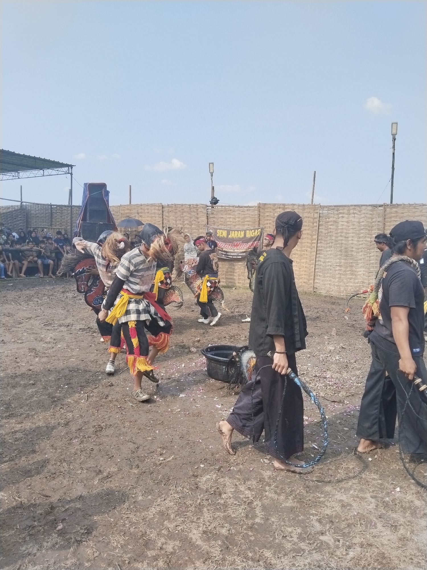 The image captures a cultural event in Kedawung, Indonesia, featuring a Jaran Kepang performance. Several individuals are dressed in traditional costumes, including woven horse-like figures, while others wear black attire and carry whips. The performance takes place in a muddy arena surrounded by a woven fence, with spectators seated in the background under a partially covered structure. A sign with the words "SENI JARAN BIGAS" is visible on the fence. Petals are scattered on the ground.