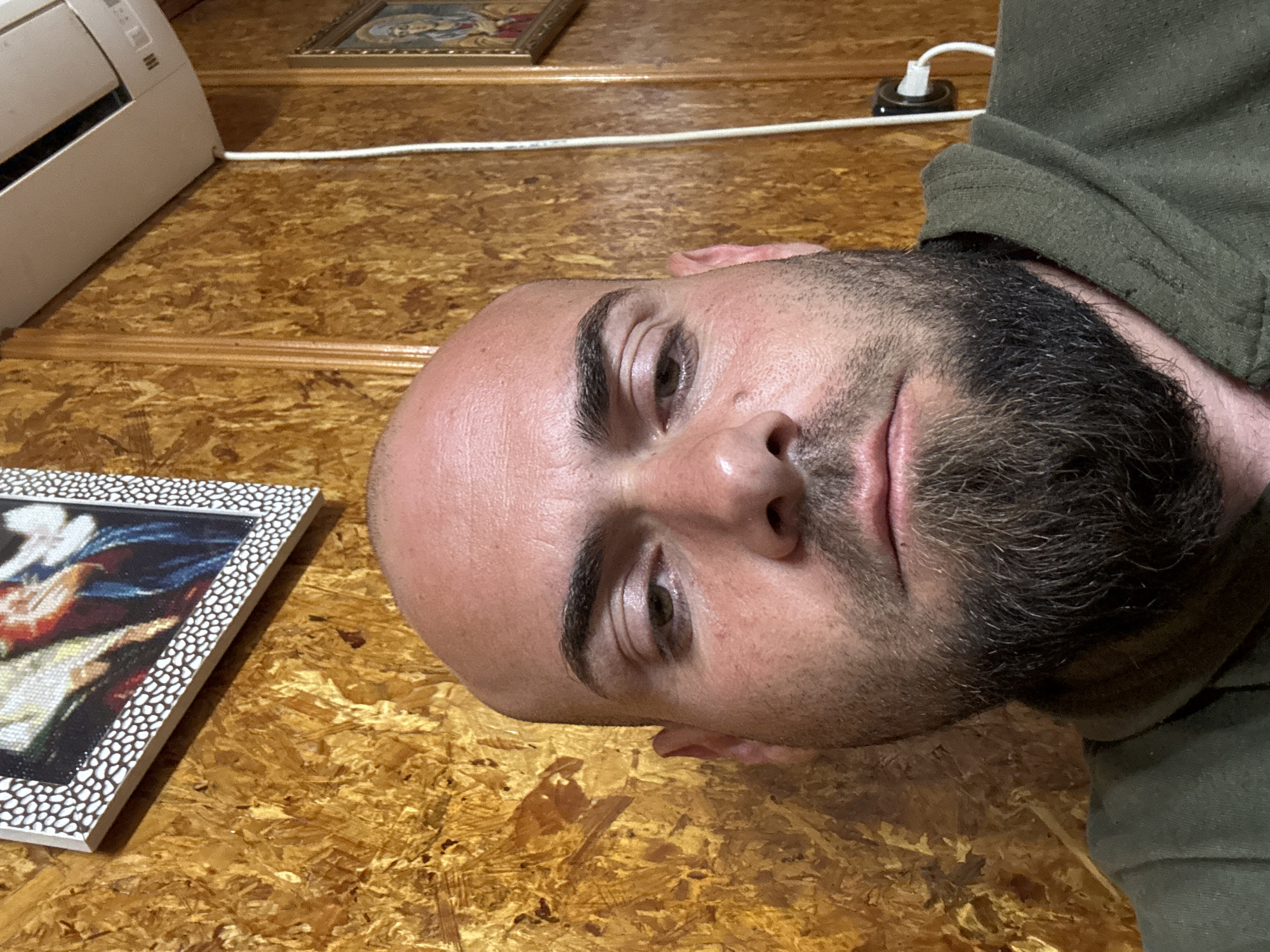 A selfie of a bald man with a dark beard, wearing a green garment. The background shows a wood-like surface, a white air conditioning unit, a framed painting or photo, and a white electrical cord with a black plug. The location in Sarnevo, Bulgaria is not discernible from the image.