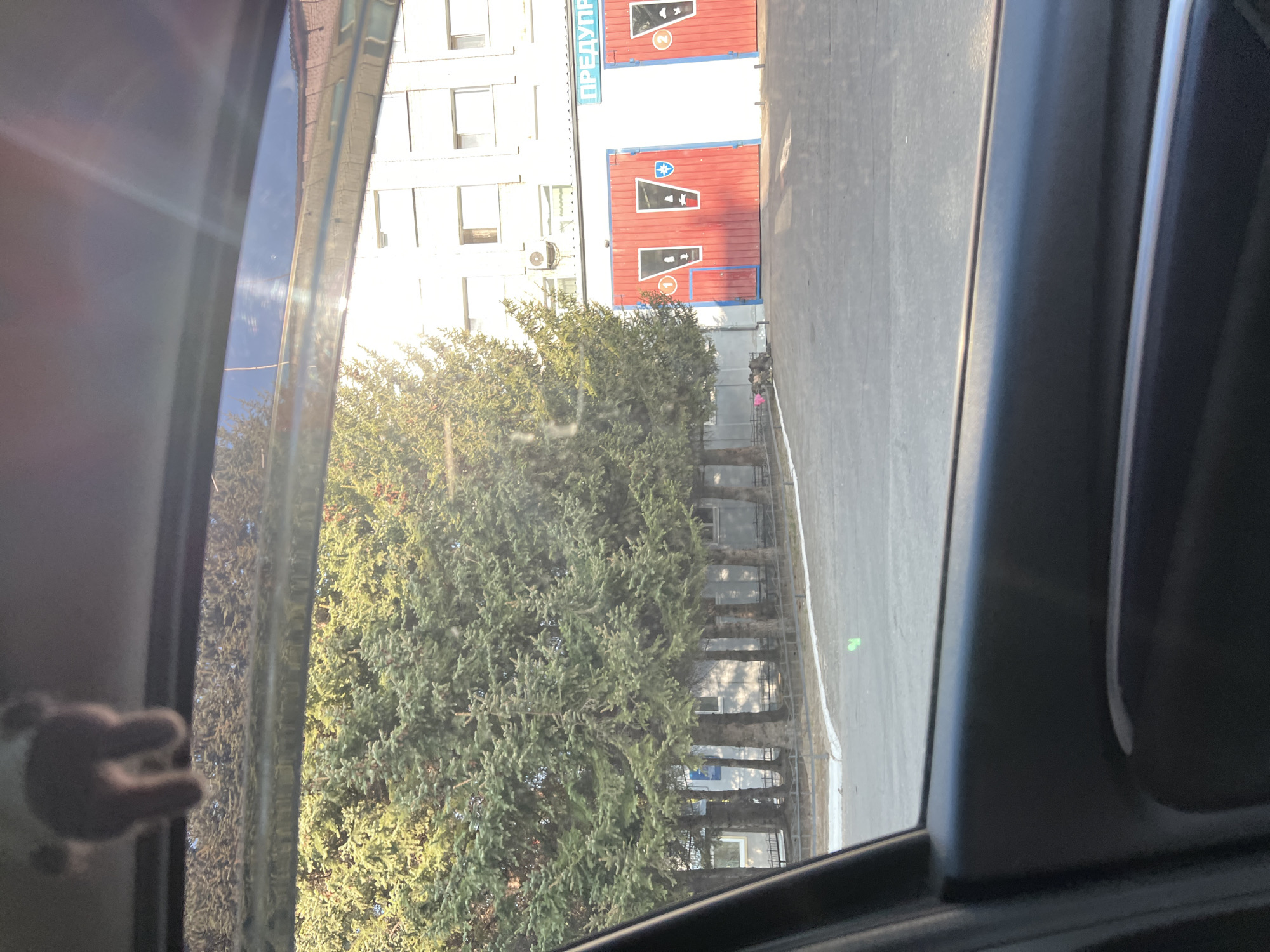 The view from inside a vehicle shows a tree-lined street alongside a multi-story building and a two-bay garage in Unknown, Unknown. The building is white with numerous rectangular windows, and the garage has red doors with painted symbols and the text "ПРЕДУПР". The trees obscure some of the details of the building behind them. The street is mostly empty with some indistinct figures. A child's hand holding a stuffed animal is visible in the lower left corner of the frame.