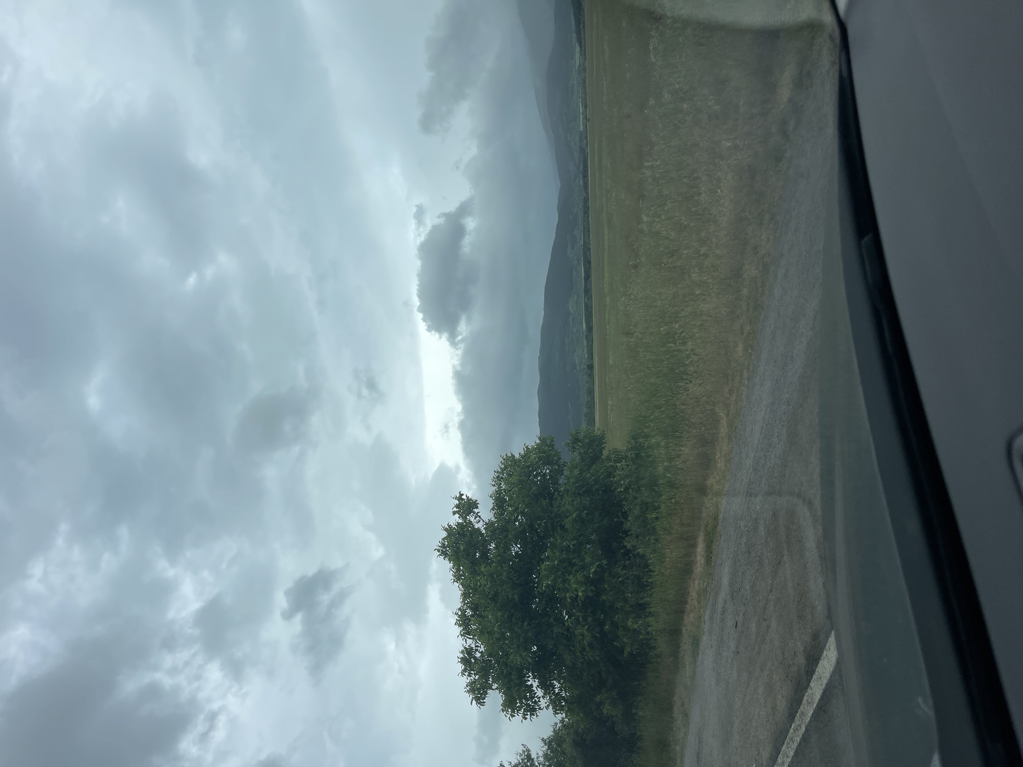 The image, taken from inside a vehicle, depicts a cloudy sky and a landscape beside a road. Mountains are visible in the distance under the low, gray cloud cover. In the foreground, a green tree is situated next to the road. The presence of streaks on the windshield suggests it is raining. The scene is possibly in Konare, Bulgaria, based on the user's claim. There are no people visible.