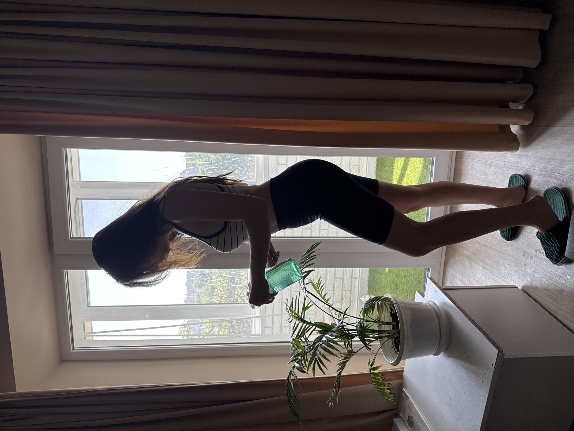 A young woman stands by a window in Kryvyi Rih, Ukraine, watering a potted plant. She is wearing a striped sports bra and black shorts. The room is brightly lit by sunlight streaming through the window. The plant is a palm tree in a white pot. There are beige curtains on either side of the window. The floor is light-colored wood.