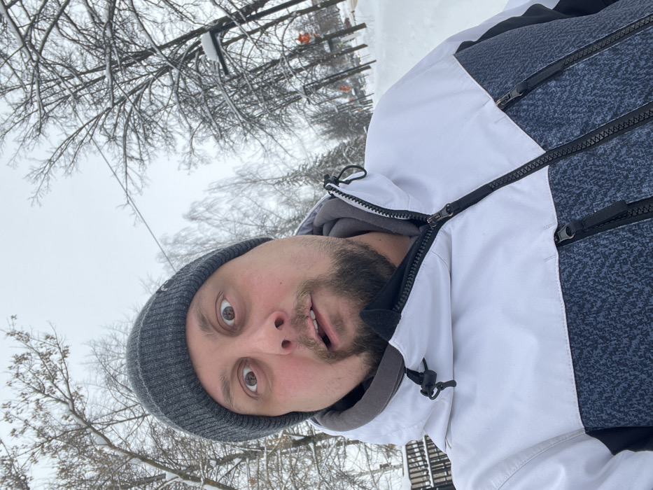 A male individual with a short beard, wearing a dark grey knit beanie and a white, grey, and black winter jacket, is depicted in a close-up selfie in the city of Chaikovsky, Russia. His eyes are wide open, eyebrows raised, and mouth slightly ajar, creating a surprised or exaggerated expression as he looks directly into the camera. The background reveals a snowy environment with numerous bare deciduous trees and some evergreen trees, all covered in white snow. The ground is entirely blanketed in snow, suggesting a park path or sidewalk. In the right background, a street lamp, power lines, and a street or road with indistinct orange objects (possibly traffic cones or signs) are partially visible. A dark, fence-like structure is discernible on the far left. The sky is overcast and grey.