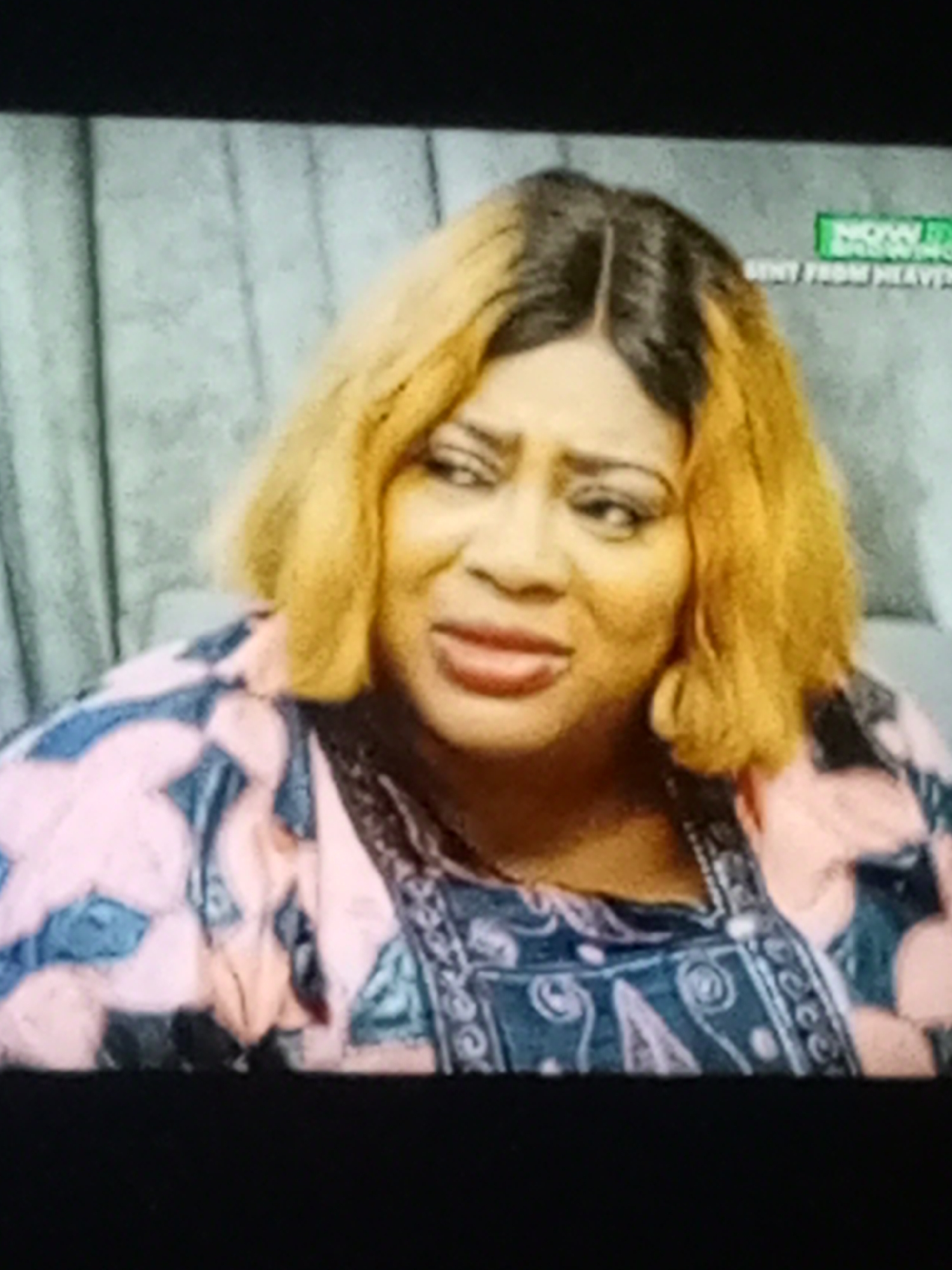 A woman with blonde hair and a patterned shirt is visible in the image. She appears to be indoors, possibly on a set. The location is unknown.