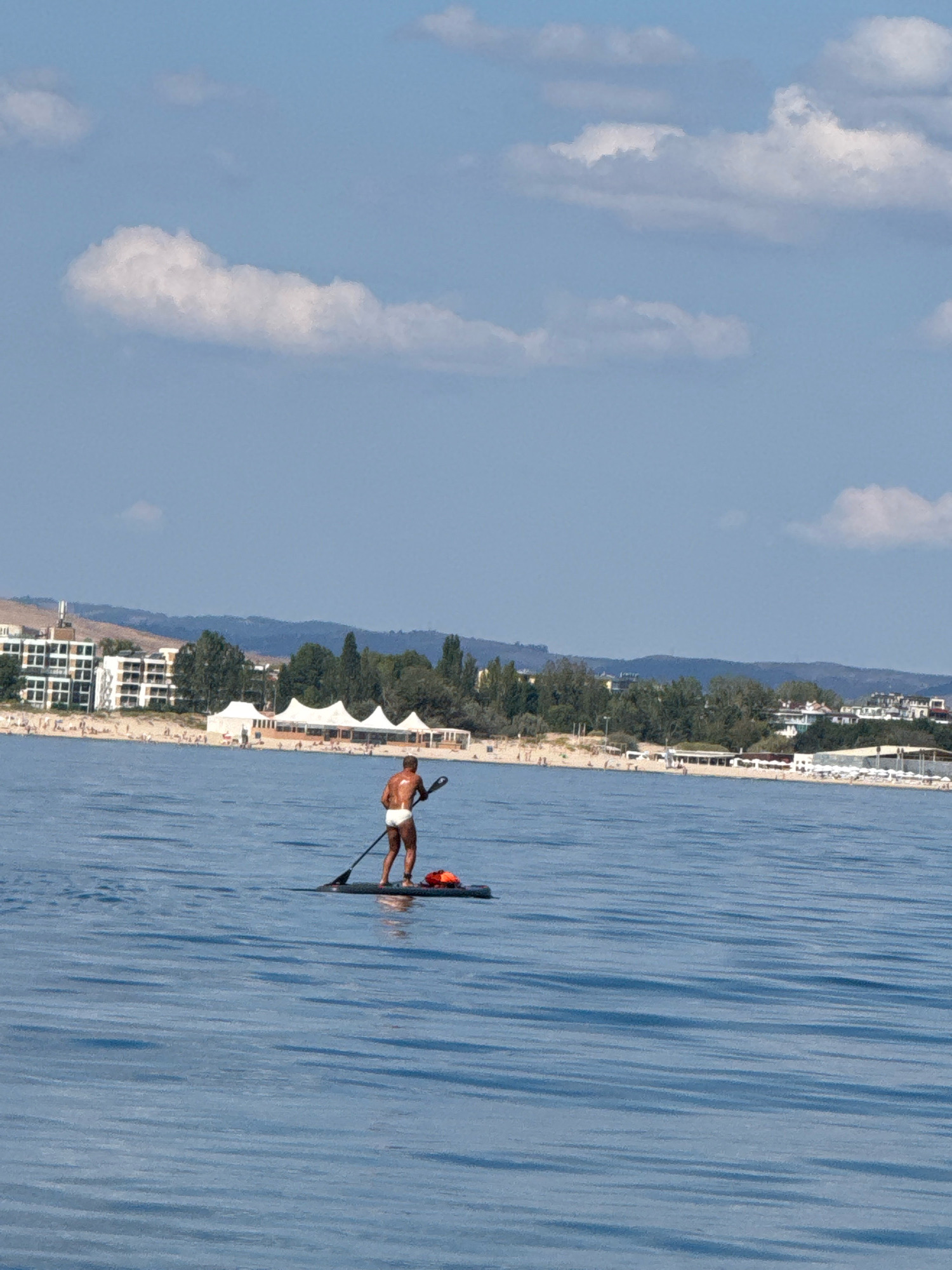 A man in white swim trunks is paddleboarding on the calm blue sea, facing away from the camera. He is standing on a dark-colored paddleboard with an orange life vest positioned behind him. In the background, a beach with buildings, white tents, trees, and people is visible along the coastline. The sky above is blue with scattered white clouds. The scene appears to be the coast of Nesebar, Bulgaria, known for its beaches and historical sites.
