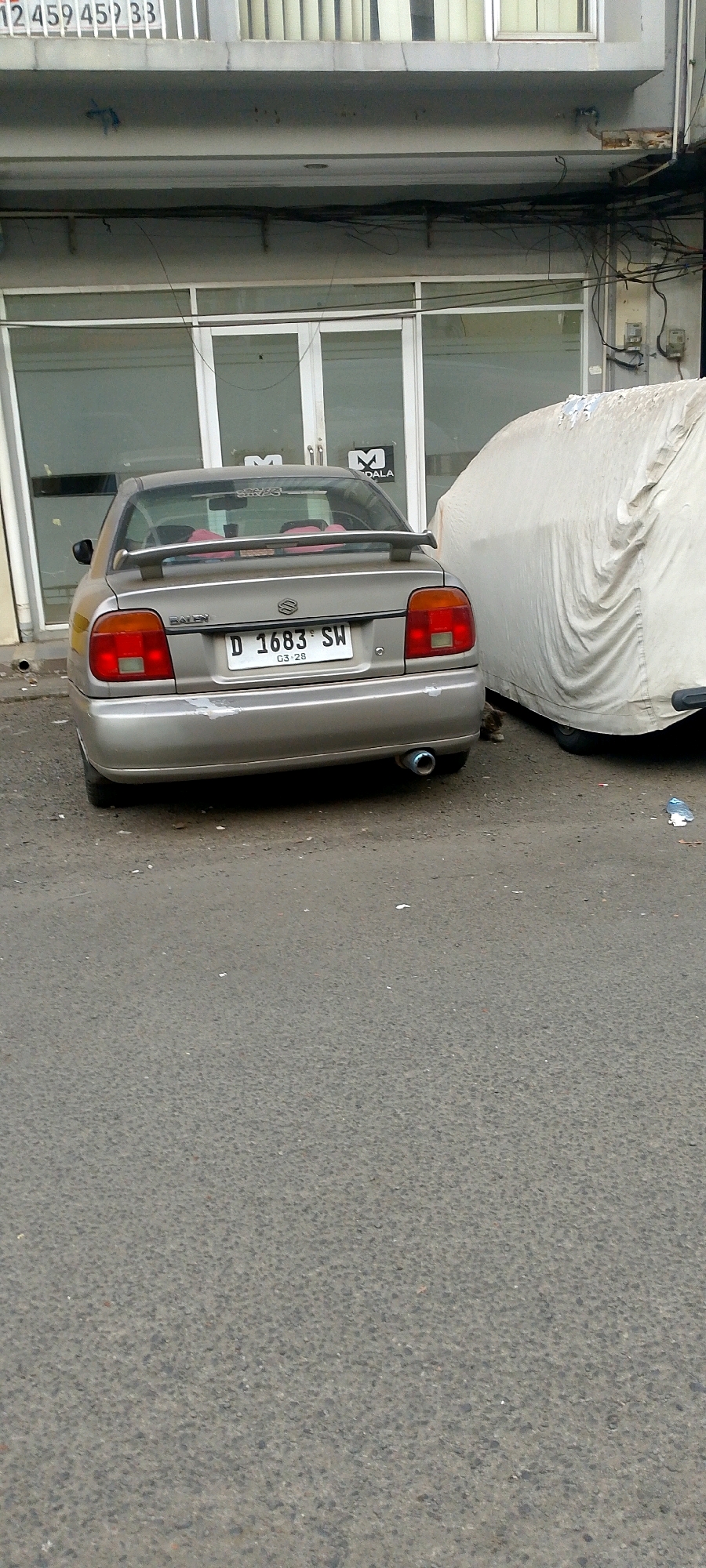 A silver Suzuki Esteem sedan is parked on an asphalt road in front of a building with large glass windows. The car is facing away from the camera and has a spoiler on the trunk. The license plate reads "D 1683 SW". To the right of the sedan, another vehicle is covered with a light-colored tarp. A small cat is partially visible between the two vehicles. The building appears to be commercial, with signage that includes the word "MALA". The road is littered with some debris. The image was taken on a cloudy day, and the overall lighting is subdued. This scene is consistent with an urban environment in Bandung City, Indonesia, given the style of the building and the vehicle's license plate format.