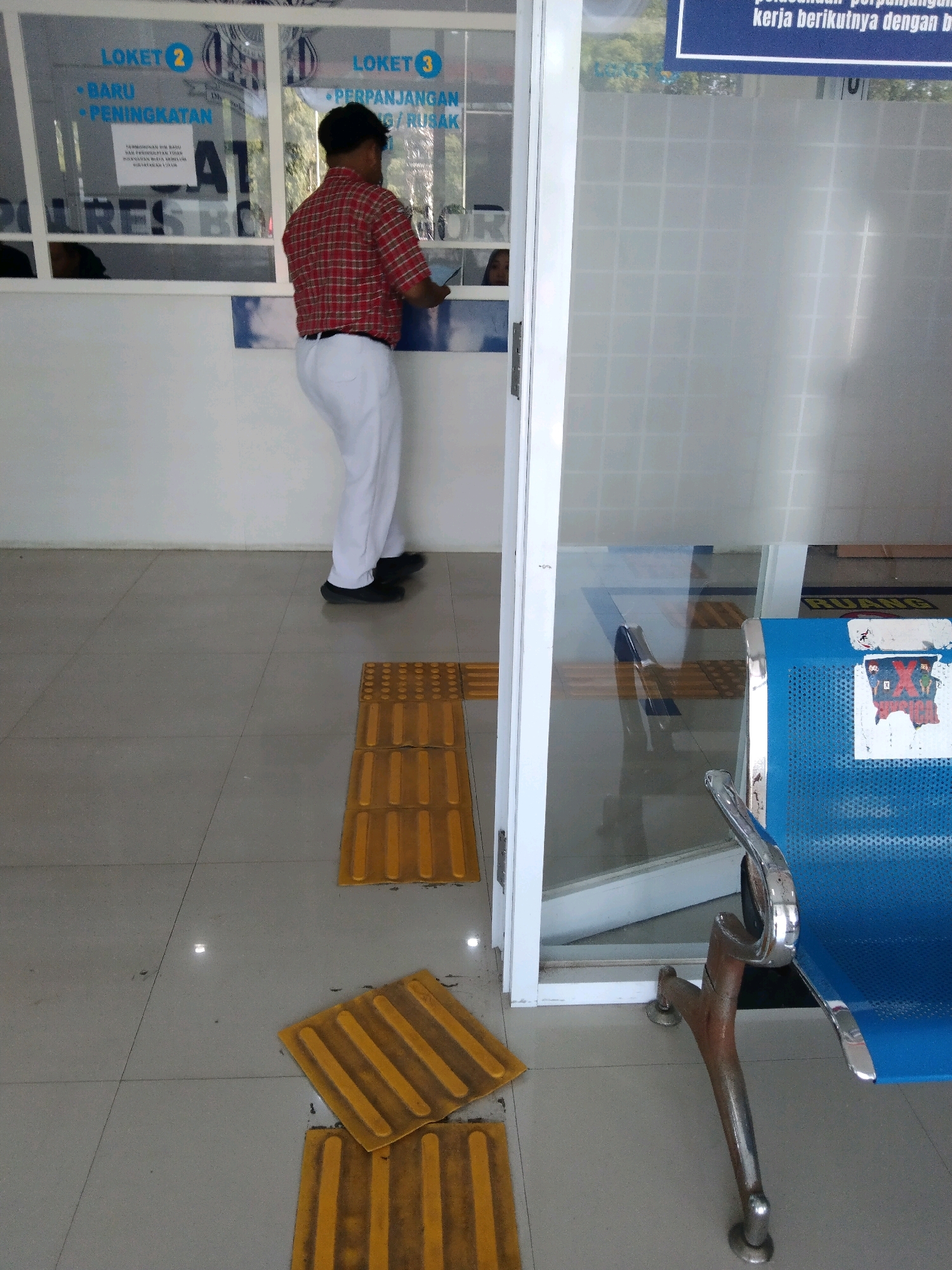 The image shows a man wearing a red and black plaid shirt and white pants standing at a counter. He is facing away from the camera and appears to be interacting with someone on the other side of the counter. The counter has signs above it that say "LOKET 2" and "LOKET 3". There are also yellow tactile paving tiles on the floor. In the background, there is a blue chair with some stickers on it. The scene appears to be an indoor public space, possibly a government office or a service center in Bojonegoro, Indonesia. The overall lighting suggests daytime.