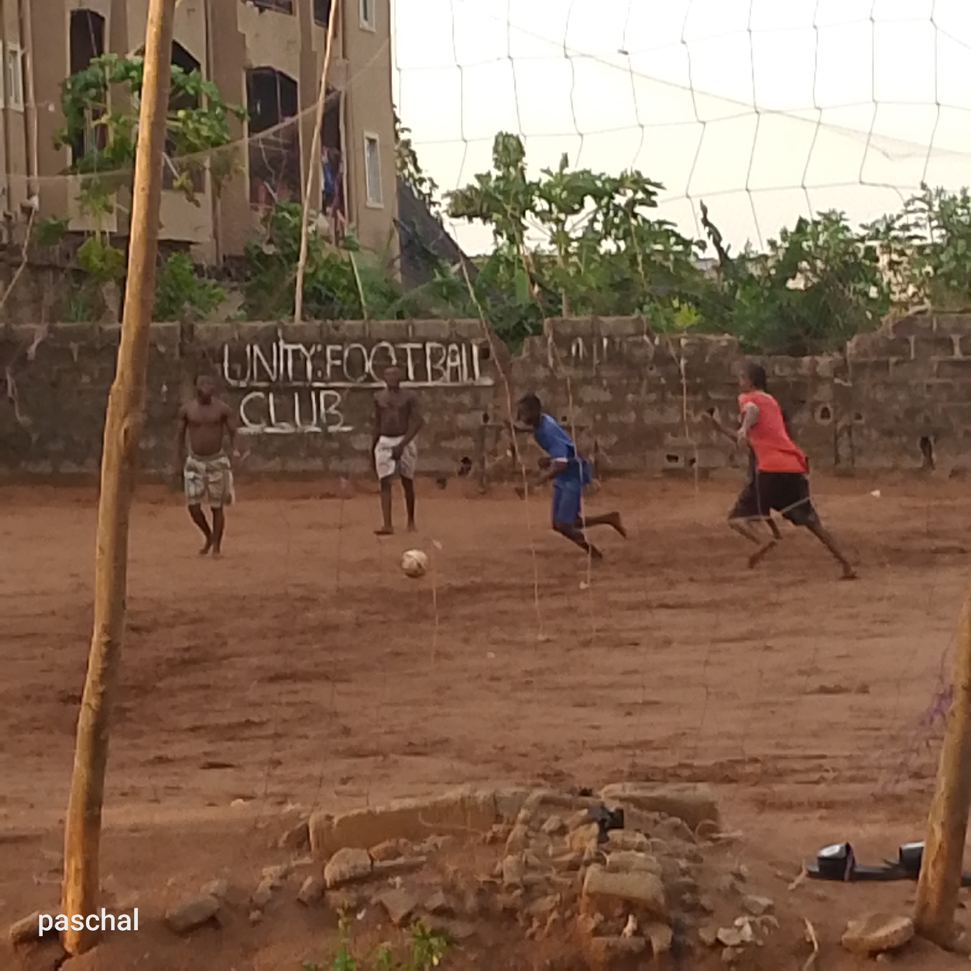 A dusty outdoor football field in Ukwu-Abwa, Nigeria, features four young men. Two men, shirtless, stand on the left of the field. One wears patterned shorts. A man in a blue shirt and shorts runs toward the ball. A man in an orange shirt and black shorts runs toward the right side of the frame. A hand-painted sign on a brick wall reads "UNITY FOOTBALL CLUB." A rough-hewn goal post and a net are in the foreground. Buildings and trees are in the background. A pair of shoes sit in the lower-right corner. The photo is labeled "paschal" in the lower-left.
