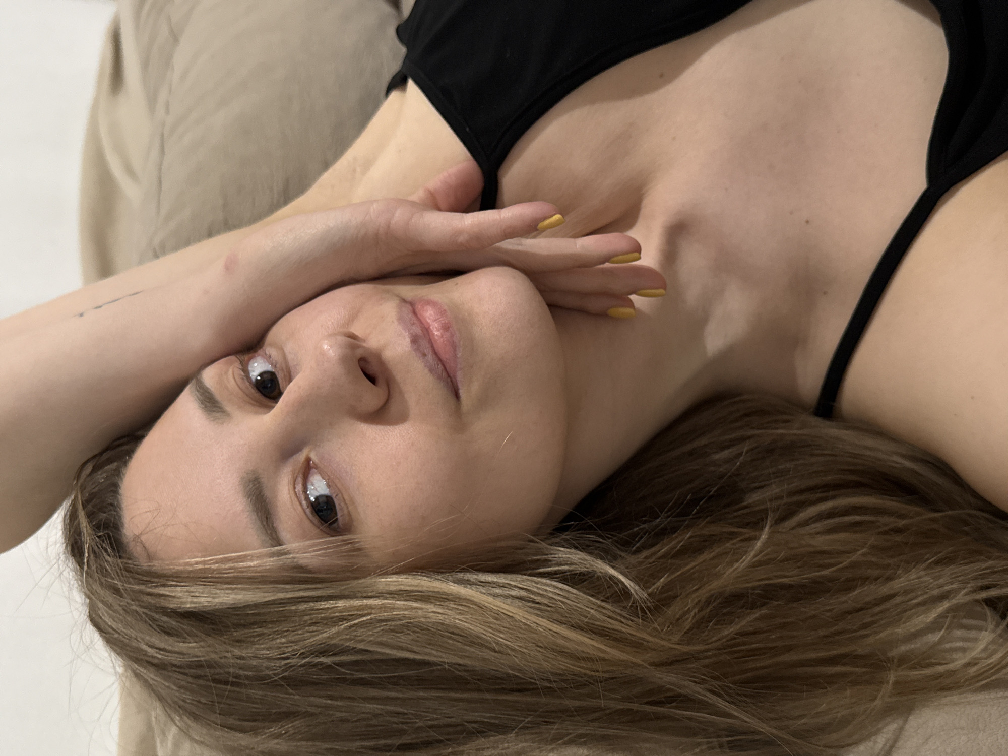A woman with blonde hair is lying down, with her head tilted towards the camera. Her hand is partially covering her face. She is wearing a black top. The background appears to be a light brown fabric or cushion. No location is identifiable in the image.