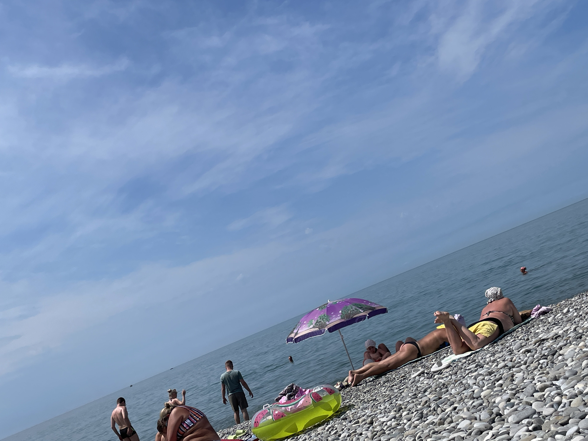 The image depicts a sunny day at a pebble beach with a clear blue sky and calm sea. Several people are present on the beach, some sunbathing on towels, others standing near the water's edge, and at least one person swimming in the sea. An umbrella provides shade for some individuals. A colorful inflatable ring is visible on the pebbles. The scene is angled, with the sea meeting the sky in the background. The people are dressed in swimwear, suggesting a recreational beach setting.