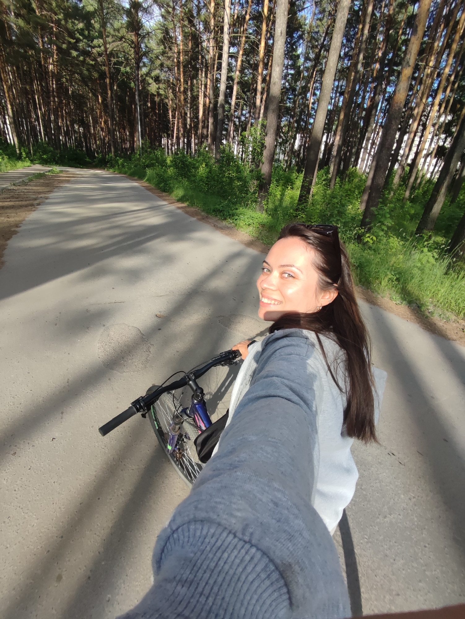 A woman is riding a bicycle on a paved path through a forest. She is wearing a light gray hooded sweatshirt and has long, dark hair. She is smiling and looking back at the camera, holding out her right arm as if to take a selfie. The path is surrounded by tall pine trees, and sunlight is filtering through the branches, casting shadows on the ground.