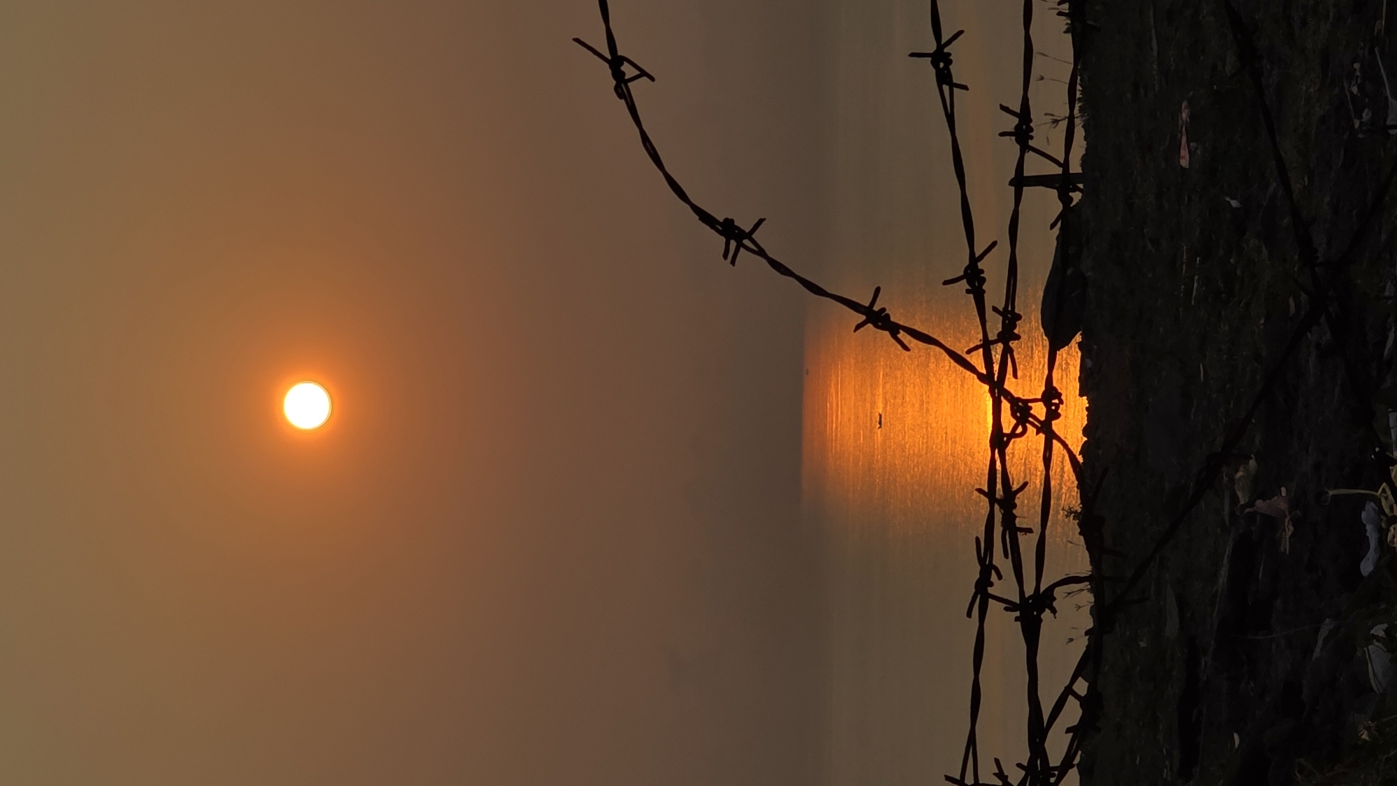 The image depicts a hazy sunset in Bac Natural Heritage Area Trang, Vietnam, with the sun appearing as a bright orb in the sky to the left. In the center of the frame, the sun's reflection creates a vertical stripe of light on the water's surface. Barbed wire runs across the foreground, partially obscuring the reflection and a dark, undefined mass, likely vegetation or a rocky outcrop, to the right. The scene has a muted, orange-brown tone throughout.