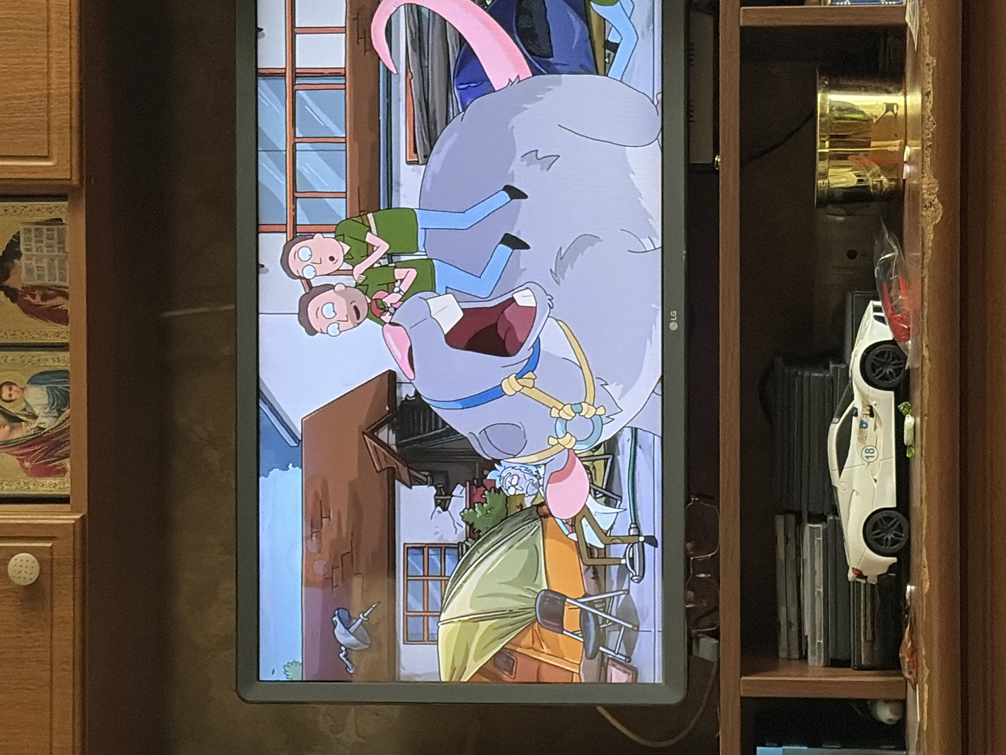 The image shows a television screen displaying an episode of Rick and Morty. Two characters, Rick and Morty, are depicted riding a giant, grey, mouse-like creature. The creature has a pink tail and is wearing a blue harness with yellow accents. Rick is in the front, and Morty is behind him. The background on the screen shows a house with a green tent and a folded chair.
Below the television, a wooden shelf holds several items. On the left side of the shelf, there is a stack of what appear to be DVDs or game cases. A white toy sports car with the number 18 on its side is placed in front of the stack. To the right of the car, there is a golden cylindrical object, possibly a container or trophy. Various small objects, including what appears to be a red toy and some white pieces, are scattered on the shelf. The overall environment suggests a living room or entertainment area.