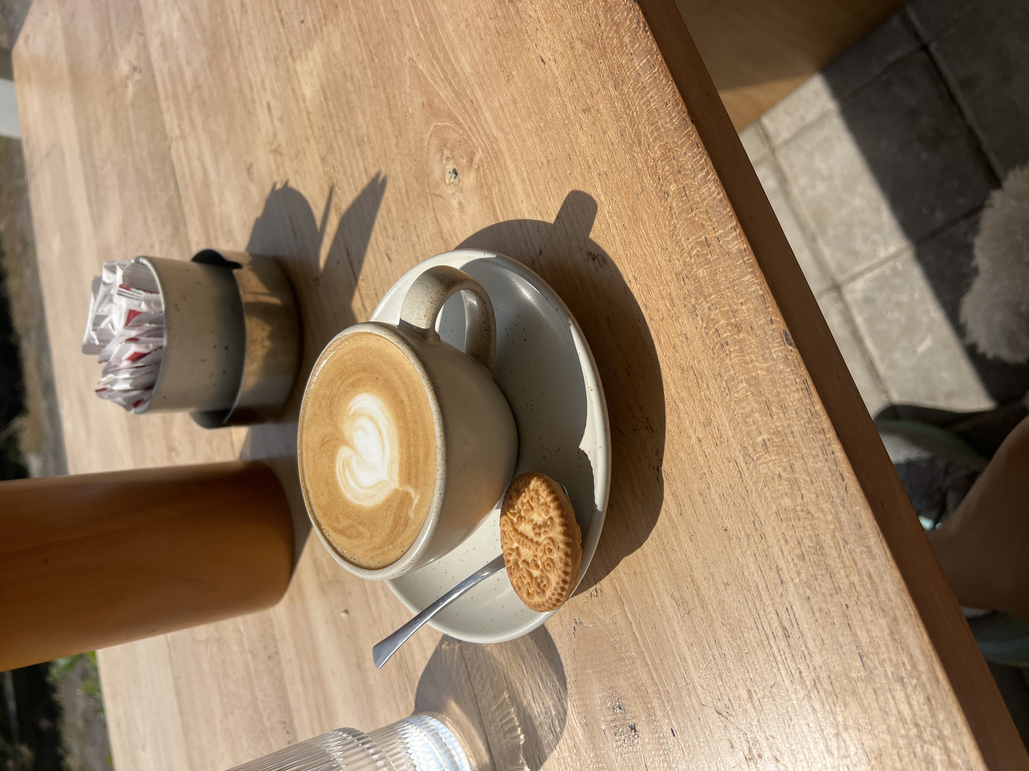 A close-up, high-angle shot shows a cappuccino with latte art in the shape of a heart, a biscuit on a spoon, and a clear glass of water on a light brown wooden table. In the background, a tall brown thermos and a small metal container filled with sugar packets are visible. The scene appears to be outdoors, possibly a cafe, with dappled sunlight casting shadows on the table and the tiled ground below. The overall impression is one of a casual outdoor cafe setting, likely in Mengwi, Indonesia, given the context.
