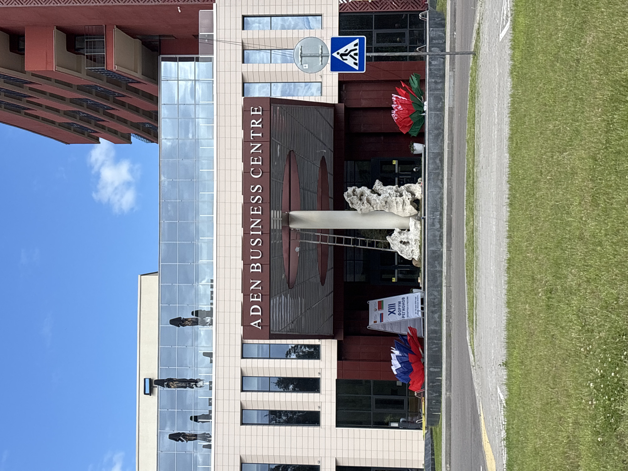 The image depicts the facade of a modern building, identified by the text "ADEN BUSINESS CENTRE". The building features a combination of cream-colored tiled walls and large glass windows, which reflect the surrounding environment and sky. To the left, a portion of a red, multi-story building with balconies is visible. A blue pedestrian crossing sign is mounted on a pole.
In front of the business center, there are decorative elements, including a large, abstract rock sculpture and two large, fan-shaped objects in red, white, and blue, resembling umbrellas or large flowers. A banner is also displayed, with visible text in Cyrillic, which reads "XIII ФОРУМ РЕГИОНОВ БЕЛАРУСИ И РОССИИ" (XIII FORUM OF REGIONS OF BELARUS AND RUSSIA). This suggests an event related to regional cooperation between Belarus and Russia is taking place or has recently concluded.
The scene appears to be outdoors, with a bright blue sky and a patch of white clouds. Adjacent to the building is a paved walkway and a strip of green grass, indicating an urban or suburban setting. The lighting suggests it is daytime, likely mid-morning or afternoon, with no strong shadows. The overall atmosphere seems neutral and business-oriented, with the decorative elements adding a touch of color and visual interest. No people are explicitly visible in the foreground or interacting with the scene, though reflections in the windows might contain them.