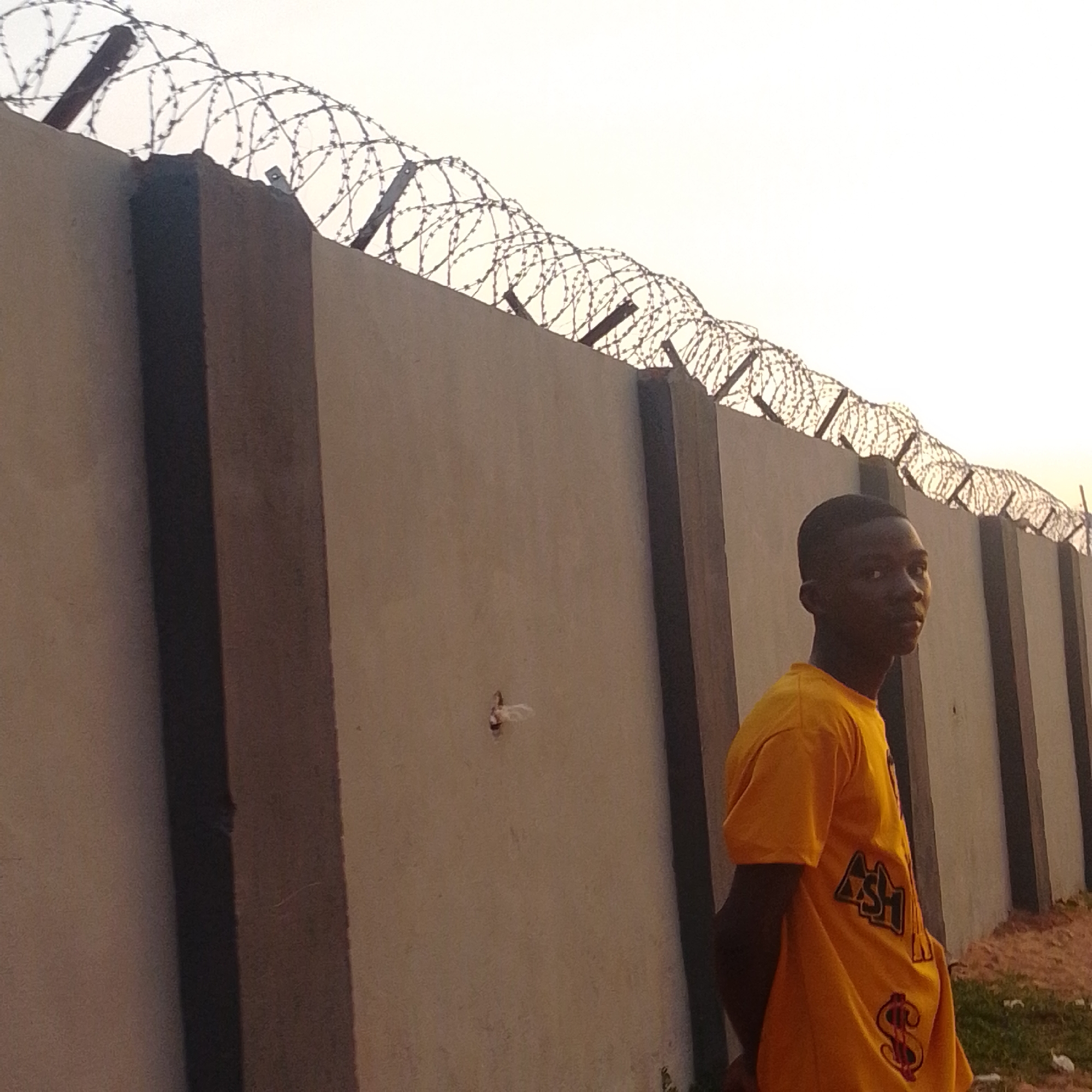 A young Black male wearing a yellow t-shirt stands next to a concrete wall topped with barbed wire. The wall is tall and stretches horizontally across the frame. The t-shirt has a graphic on it that includes the letters "ASH" in a black triangle with a dollar sign below it. The sky is bright and appears to be overcast or at dusk. The ground in front of the wall is dirt and some patchy grass. The scene is likely outdoors.