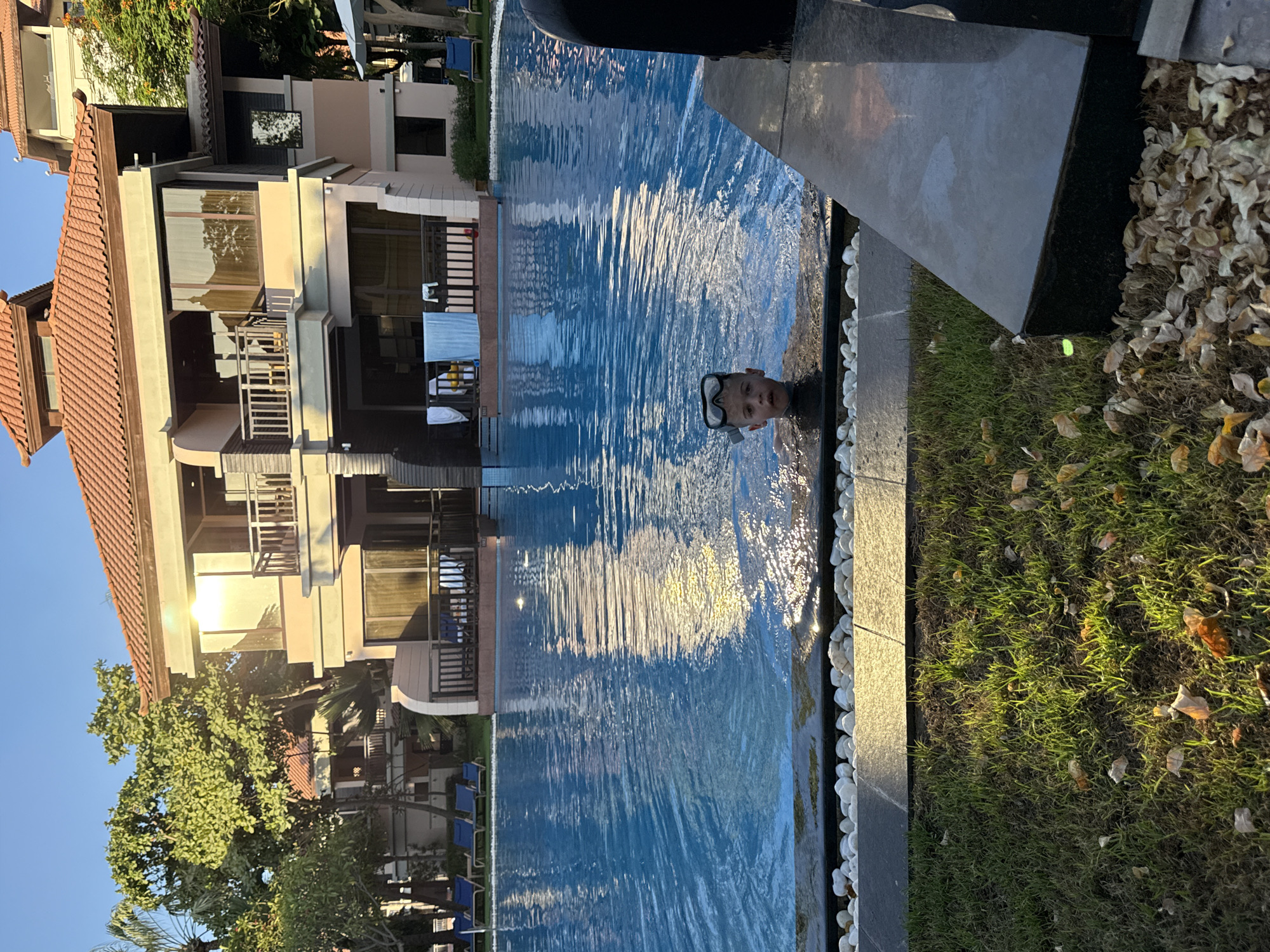 A young boy wearing a snorkel mask and breathing tube is in a swimming pool. The pool appears to be an infinity pool, with the water flowing over the edge. The boy is looking directly at the camera with a slight smile. The water is a vibrant blue and reflects the sunlight.
Behind the boy and to the left is a multi-story building with a tiled roof and balconies. The building appears to be a hotel or resort. Green trees and foliage are visible around the building. In the foreground, to the right of the pool, is a grassy area with fallen leaves.
The scene suggests a sunny day, likely in the late afternoon given the warm light reflecting off the building and the water. The overall mood is relaxed and happy, with the boy enjoying a swim. The location context of Dubai, United Arab Emirates, suggests a warm climate. There is no visible text in the image.
