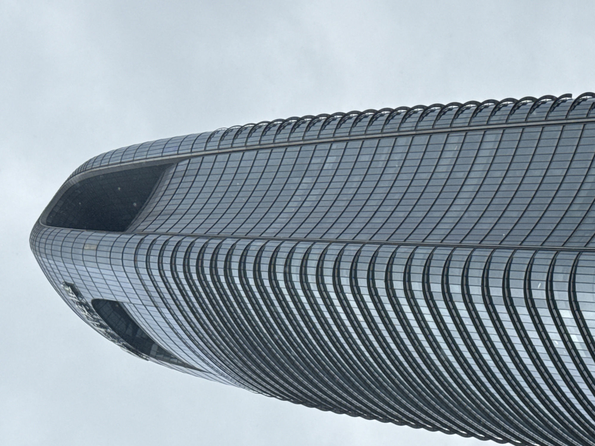 A low-angle, full-shot photograph shows the top section of a modern, elliptical skyscraper in the Haizhu District, China. The building's facade is covered in curved, reflective glass panels that create a shimmering effect. The top of the skyscraper features a dramatic, open-air cutout that resembles a crescent moon, with a horizontal element visible within the opening, possibly a walkway or structural component. The logo of the building, appearing to be "PICC" in large, stylized letters, is visible within the cutout area. The sky is overcast, with a pale gray hue, indicating a cloudy day. The building's structure appears smooth and seamless, with vertical dark lines delineating sections of the glass. The lower portion of the building is not visible in the frame.