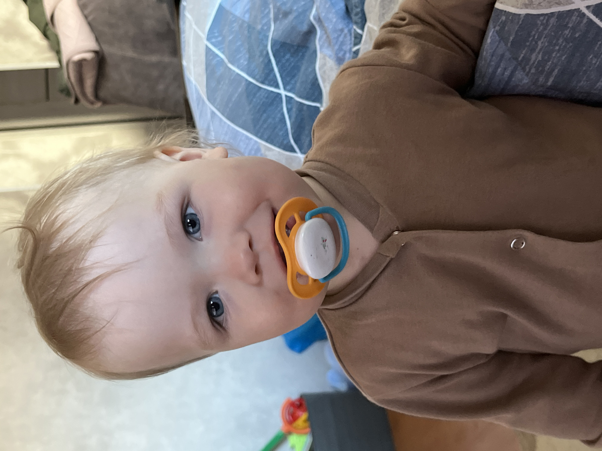 A baby with light blonde hair and blue eyes is looking up at the camera, holding a pacifier in their mouth. The pacifier has an orange base with a blue ring and a white center. The baby is wearing a brown long-sleeved onesie. The background includes a blue and white patterned pillow, a gray upholstered item, and glimpses of a colorful toy on a light-colored floor. The location appears to be indoors. I am unable to provide a more specific location in the city of Chaikovsky, Russia, as there is no identifiable information within the image to suggest that.