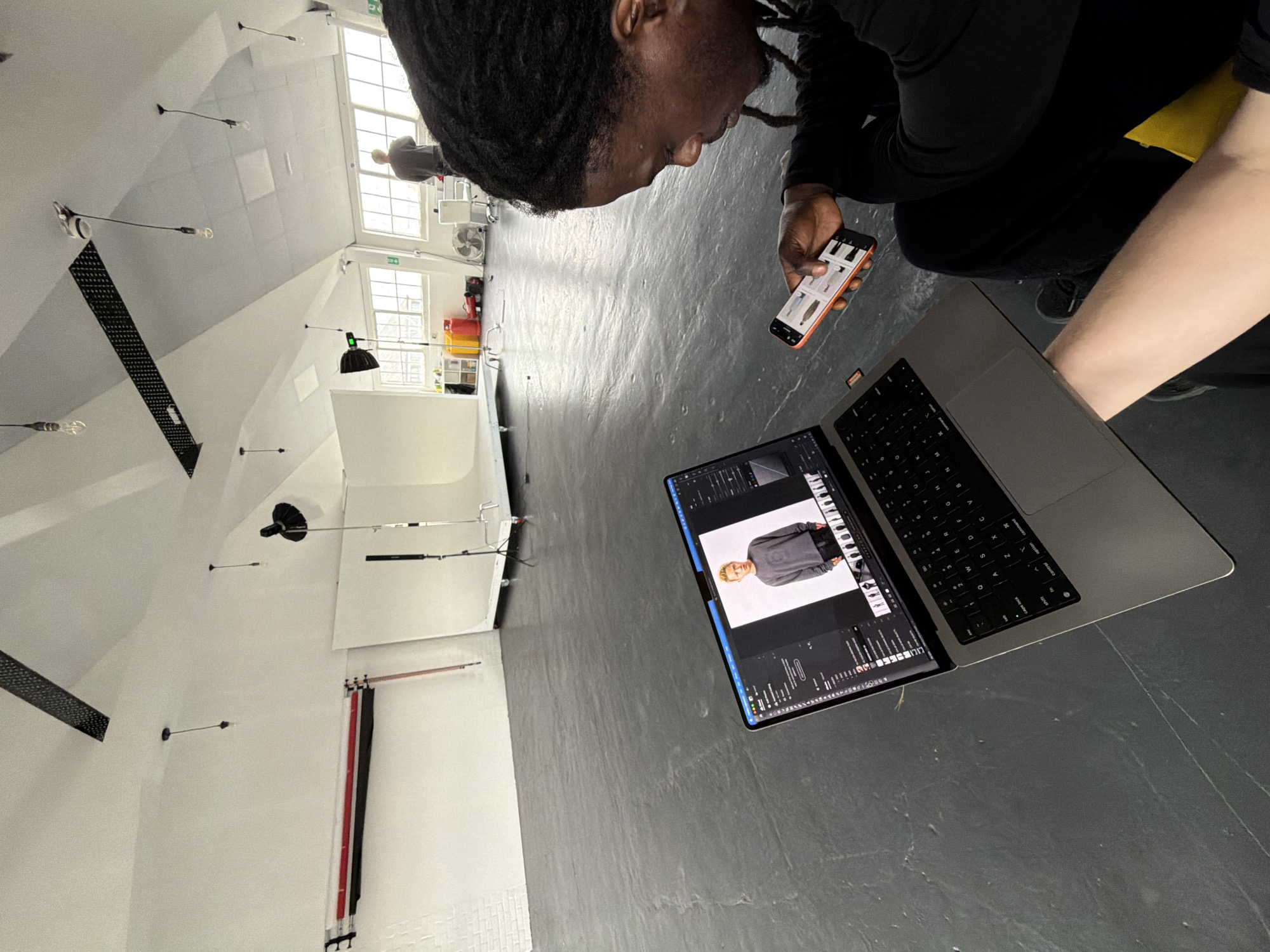 An indoor photography studio setting in London, UK, shows a man with dark hair, possibly dreadlocks, using a mobile phone while looking at a laptop screen. The laptop displays an image of a model. The floor is a grey, semi-glossy surface. Background features include studio equipment and lighting. A second person can be seen faintly in the background through the window.