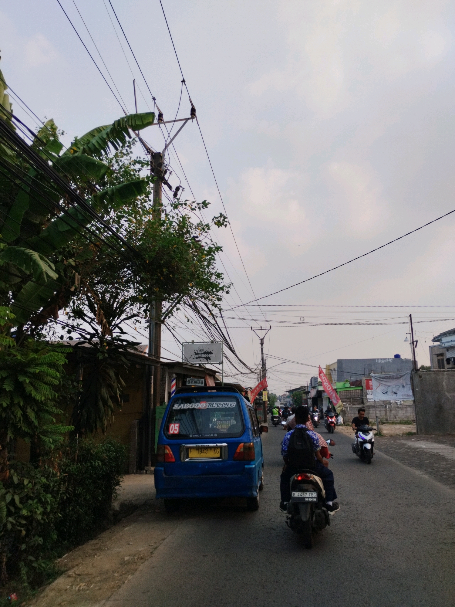The image depicts a street scene in Bojong Gede, Indonesia. A blue van is parked on the right side of the road, facing away from the camera. Its license plate is visible and reads "F 1943 KG". On the rear of the van, text is visible, including "SABODO TEUNGUL". To the left of the van, a barber pole stands vertically.
Several motorcycles are on the road. A person is riding a motorcycle directly in front of the van, their back to the camera, wearing a backpack. Another motorcycle is approaching from the opposite direction, ridden by a person wearing a black shirt and helmet. Further down the road, other motorcycles and people are visible.
The environment includes a road, sidewalks, and buildings. Lush green foliage, including banana trees, is present on the left side of the image, with dense vegetation bordering the road. Overhead, numerous electrical wires are strung across poles, typical of the area. The sky is overcast. The overall scene suggests a typical day on a street in Bojong Gede.
