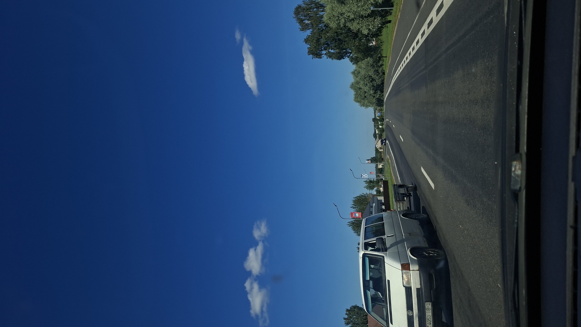 A white Volkswagen van is traveling on a highway in Myadzyel, Belarus, pulling a small trailer. The van is in the left lane, and there is a single white lane line separating it from the right lane. The road is paved and appears to be in good condition. The sky is a clear blue with a few white clouds. Trees and houses are visible on either side of the road. There is another car in the distance, also traveling on the highway. The scene appears to be from the perspective of someone inside another vehicle, looking forward.