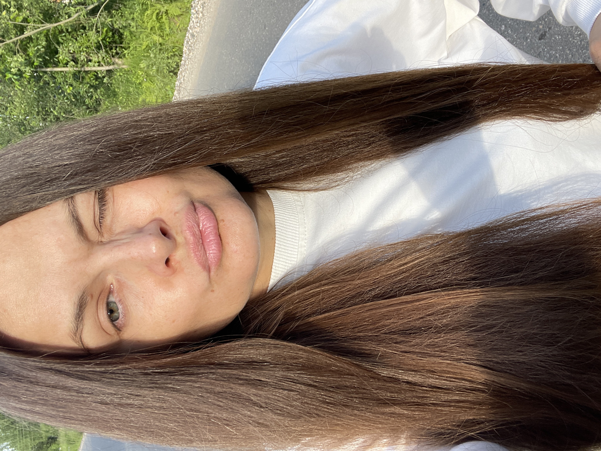 In an outdoor, daylight scene, a woman with long, dark brown hair winks at the camera. She is wearing a white crewneck sweatshirt. Her lips are pursed. The background shows an asphalt road and lush green trees. This setting appears to be a rural or suburban area on the outskirts of a city, consistent with the outskirts of Заречный, Russia.