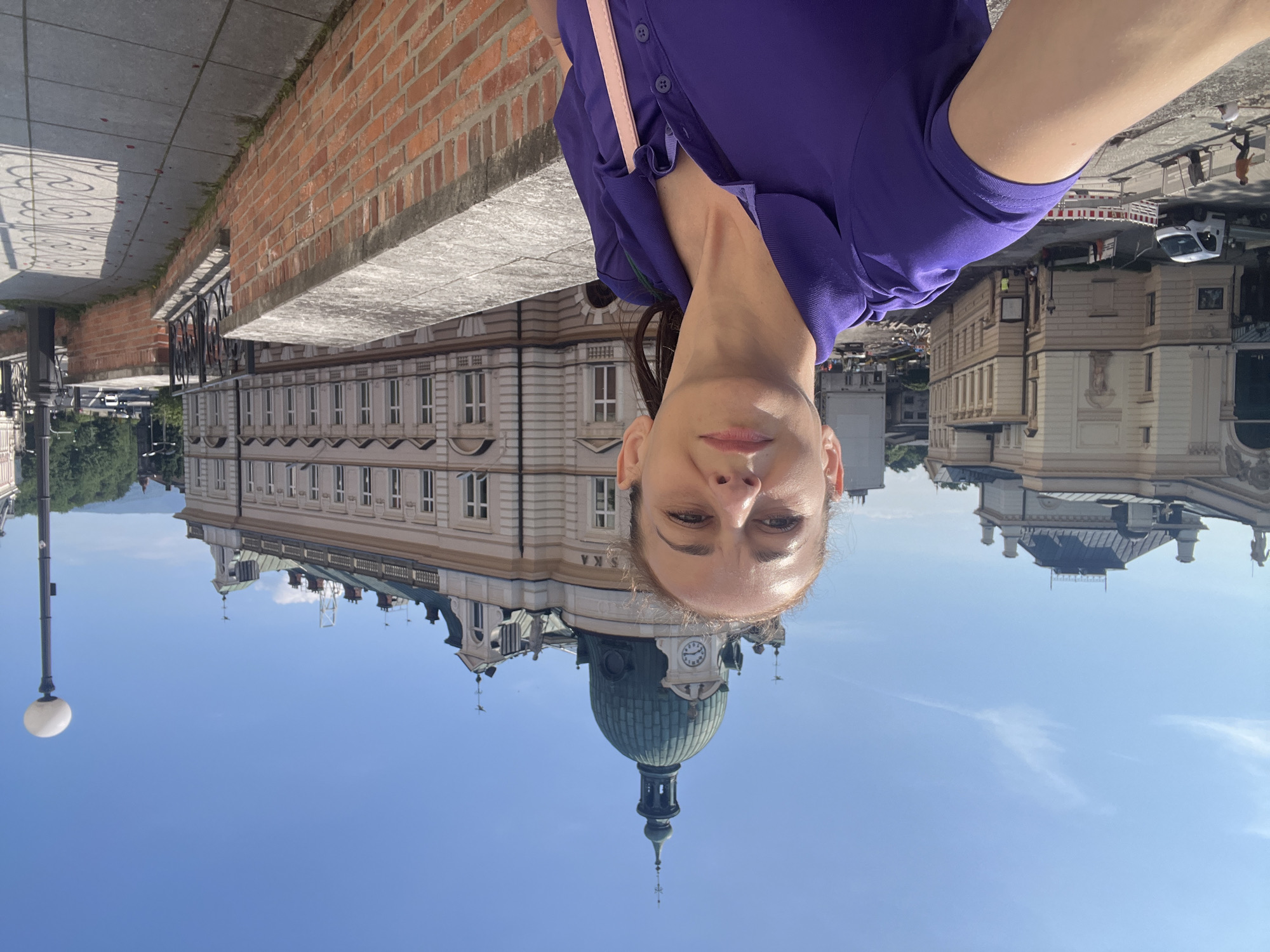 In a selfie taken outdoors, a woman with brown hair pulled back in a ponytail stands in the foreground, wearing a purple collared shirt. She is looking directly at the camera. Behind her, a large building with a green dome and a clock tower dominates the background. The building has multiple windows and decorative architectural elements. To the left of the dome building, another building with classical architecture is visible. A brick wall runs along the right side of the image, with a sidewalk next to it. The sky is clear and blue. Construction barriers and workers are visible in the distance on the left side of the frame, suggesting ongoing work in the area.