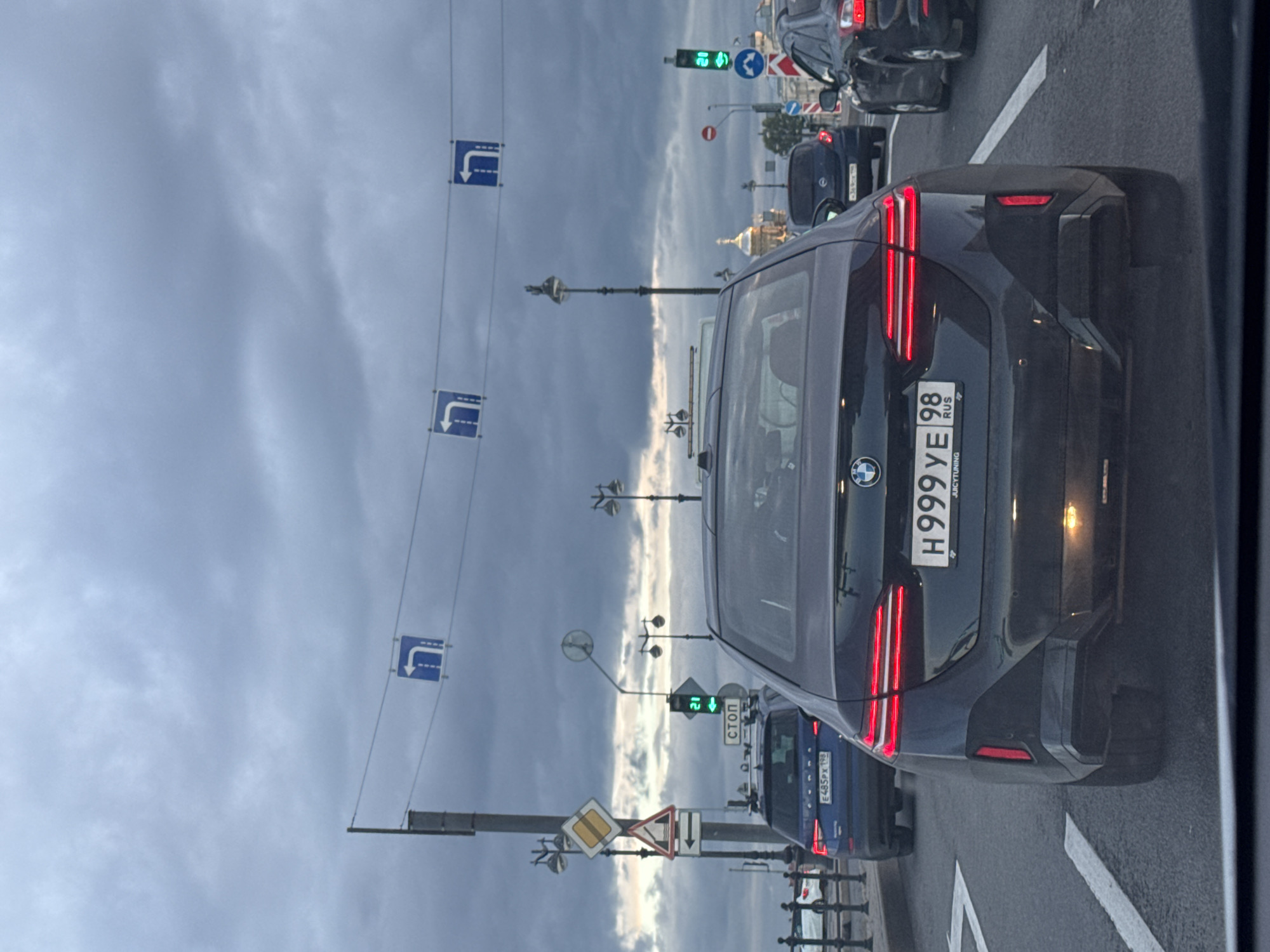 A dark, overcast day in Saint Petersburg, Russia, as evidenced by the grey, cloudy sky and the ambient light. The image is taken from inside a vehicle, looking forward at traffic on a city street. The primary subject is the rear of a black BMW sedan with its taillights illuminated. The license plate reads "H999YE 98 RUS". To the left of the BMW is a blue Skoda SUV, its license plate is partially visible as "E485PX 198". In the distance, other vehicles and streetlights are visible. Traffic signals, including a green light indicating "GO" and other directional signs are present. Overhead, blue and white directional traffic signs with white arrows indicating left turns are suspended over the roadway. Road markings, including white lane dividers, are visible on the asphalt. The architectural style of buildings in the far background suggests an urban environment, possibly near historical structures, common in Saint Petersburg.