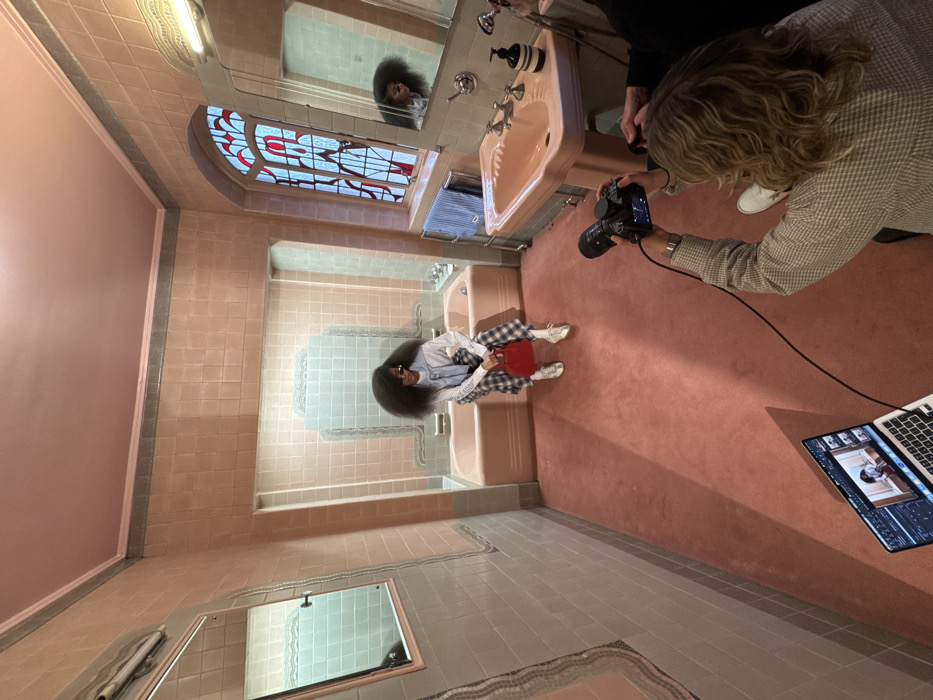 A bathroom setting, with a woman sitting on the edge of a tub, holding a small red purse. She has a voluminous hairstyle and wears sunglasses, a white shirt, and plaid pants. Another woman is taking pictures with a professional camera; a laptop on the floor displays captured images. The bathroom features vintage pink tiles, a stained-glass window, and a pink sink. The photograph appears to be a behind-the-scenes shot of a fashion photoshoot.