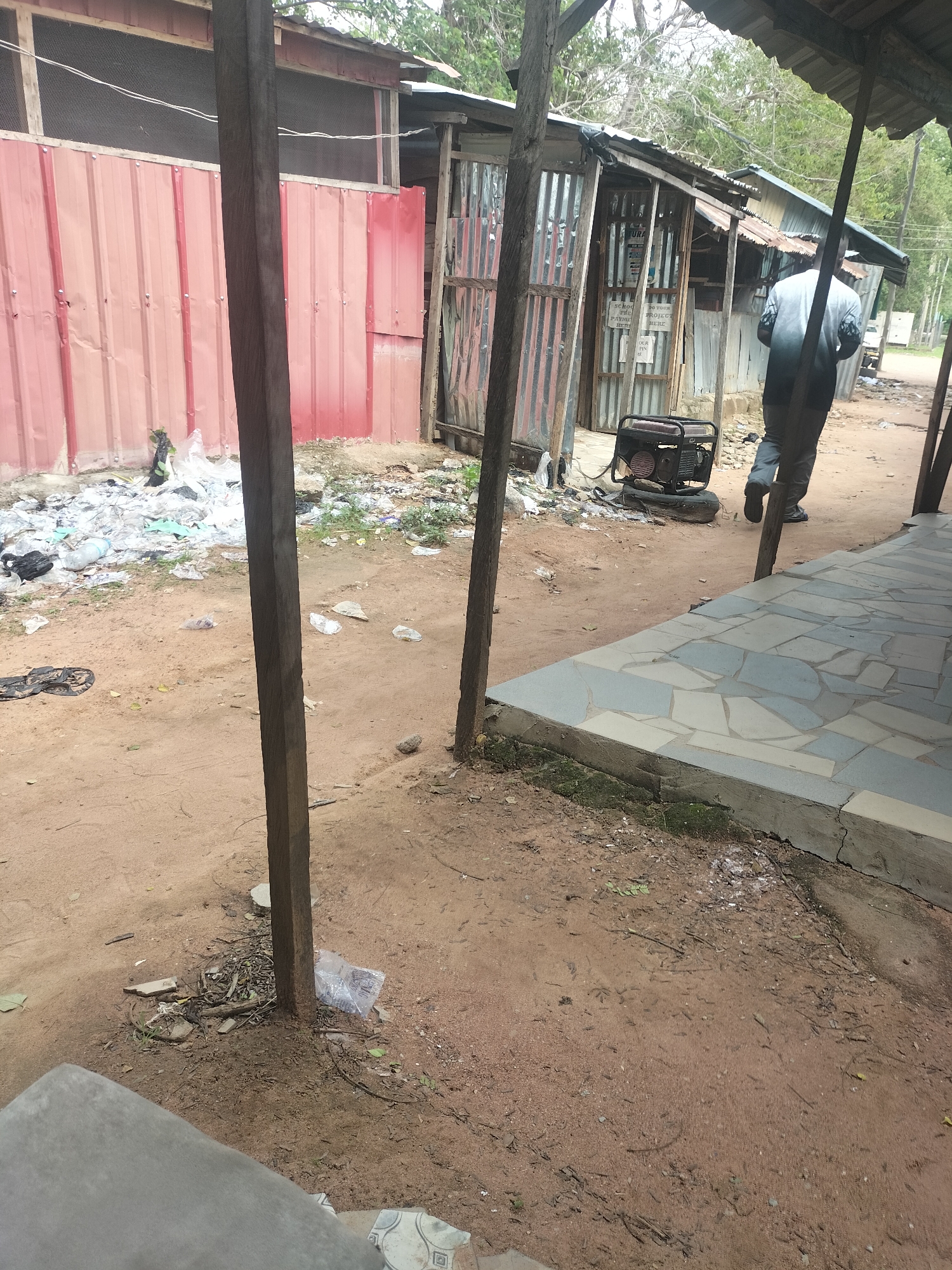The image depicts an outdoor scene in Jalingo, Nigeria. In the foreground, a sandy area is visible with some scattered debris, including plastic bags and leaves. Two wooden posts are prominent, supporting a simple roof structure on the right side. A tiled walkway runs along the right edge of the frame.
In the mid-ground, a red corrugated metal building stands on the left, partially obscured by the wooden post. Adjacent to this building is a small structure constructed with corrugated metal and wooden supports, showing signs of disrepair. A portable generator is situated in front of this structure. A person, seen from behind, is walking away from the camera towards the right, wearing a dark top with a gradient design and dark pants. Further back, a dirt road is visible with some trees and vegetation lining the sides. The overall environment appears somewhat unkempt, with visible trash accumulation near the buildings.