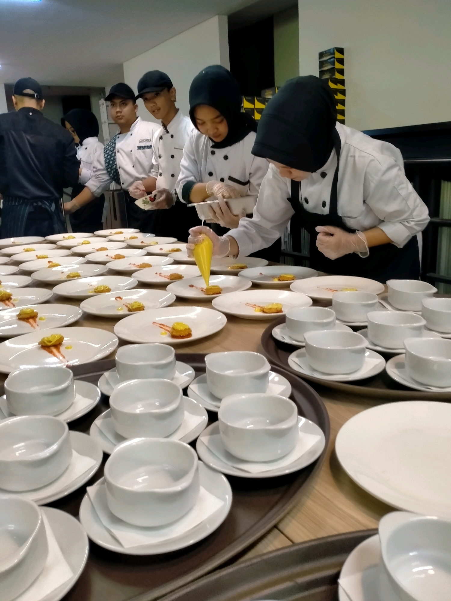 A group of culinary students are preparing appetizers in a large kitchen. In the foreground, an array of small, white appetizer plates are neatly arranged on a wooden table. Each plate features a single bite-sized appetizer, often adorned with a dollop of yellow sauce and a drizzle of red sauce. Several small, white bowls sit on saucers, some stacked on metal trays, suggesting they are for soup or a side dish. In the background, five individuals, dressed in white chef's coats and aprons, stand behind a table laden with more plates. One student, wearing a black headscarf, is in the process of piping yellow sauce onto a plate with a piping bag. Another student, also in a headscarf, holds a small plate and appears to be adding a garnish. Three male students, two wearing baseball caps and one in a black headscarf, stand to their left, observing the preparation or waiting for their turn. The overall impression is one of focused activity in a professional culinary setting, likely a training facility or a restaurant kitchen during a busy service period. The image appears to have been taken in a kitchen located in Batu, Indonesia, a city known for its culinary schools and vibrant food scene.