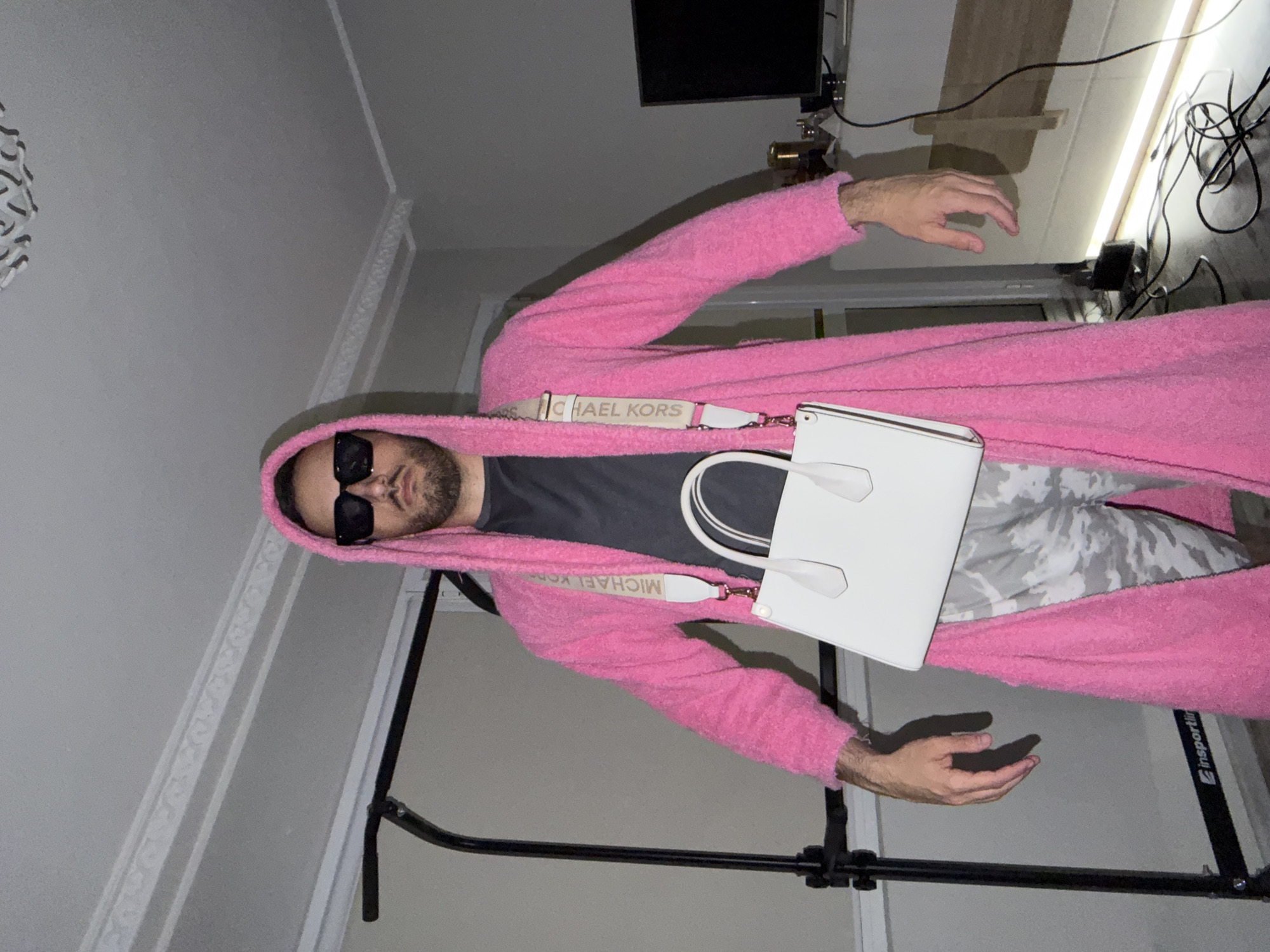 A man wearing a pink hooded robe, sunglasses, a gray t-shirt, and camo pants is holding a white purse with a "Michael Kors" strap. The background includes part of a photography light stand and household items. There's no information to suggest that the location is Plovdiv, Bulgaria.